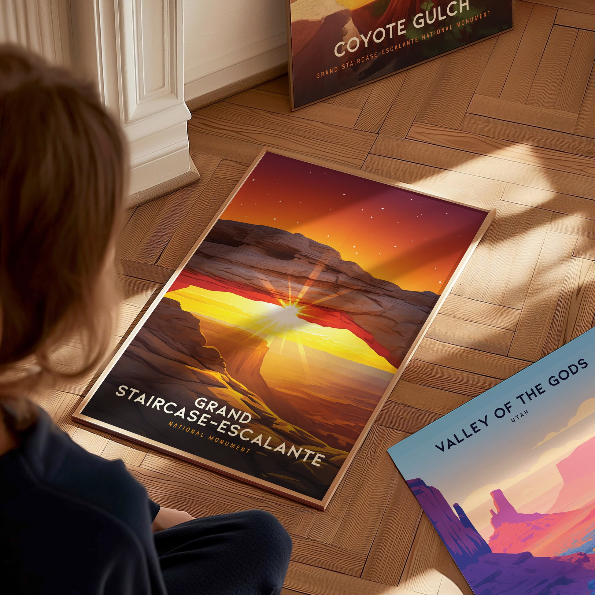 A person sits on a wooden floor looking at a Grand Staircase–Escalante poster, showcasing a vivid sunset arch scene. Nearby, other posters are partially visible.