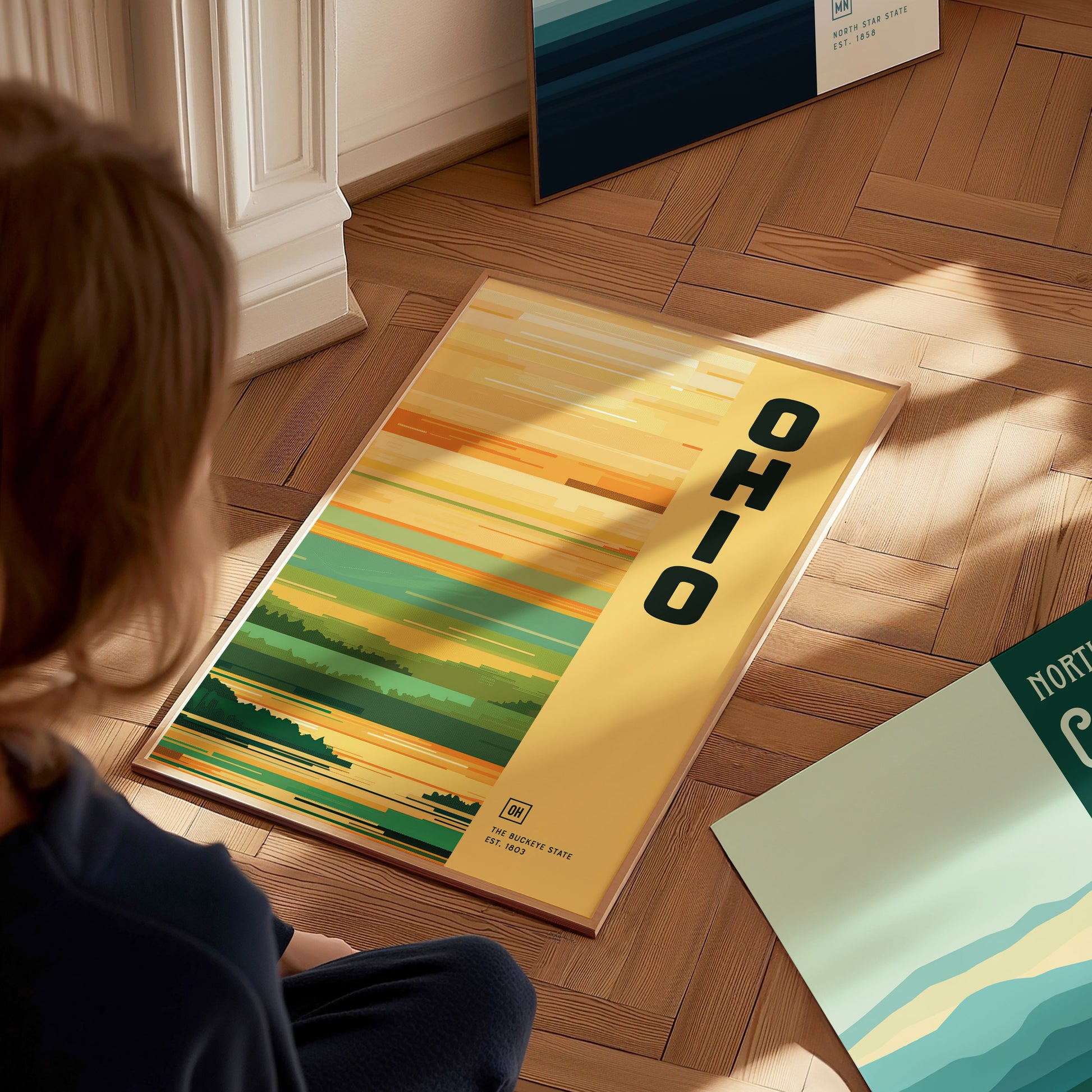 A person looks at a minimalist Ohio poster on a wooden floor, featuring colorful horizontal stripes and the word OHIO in bold. Another poster is partially visible nearby.