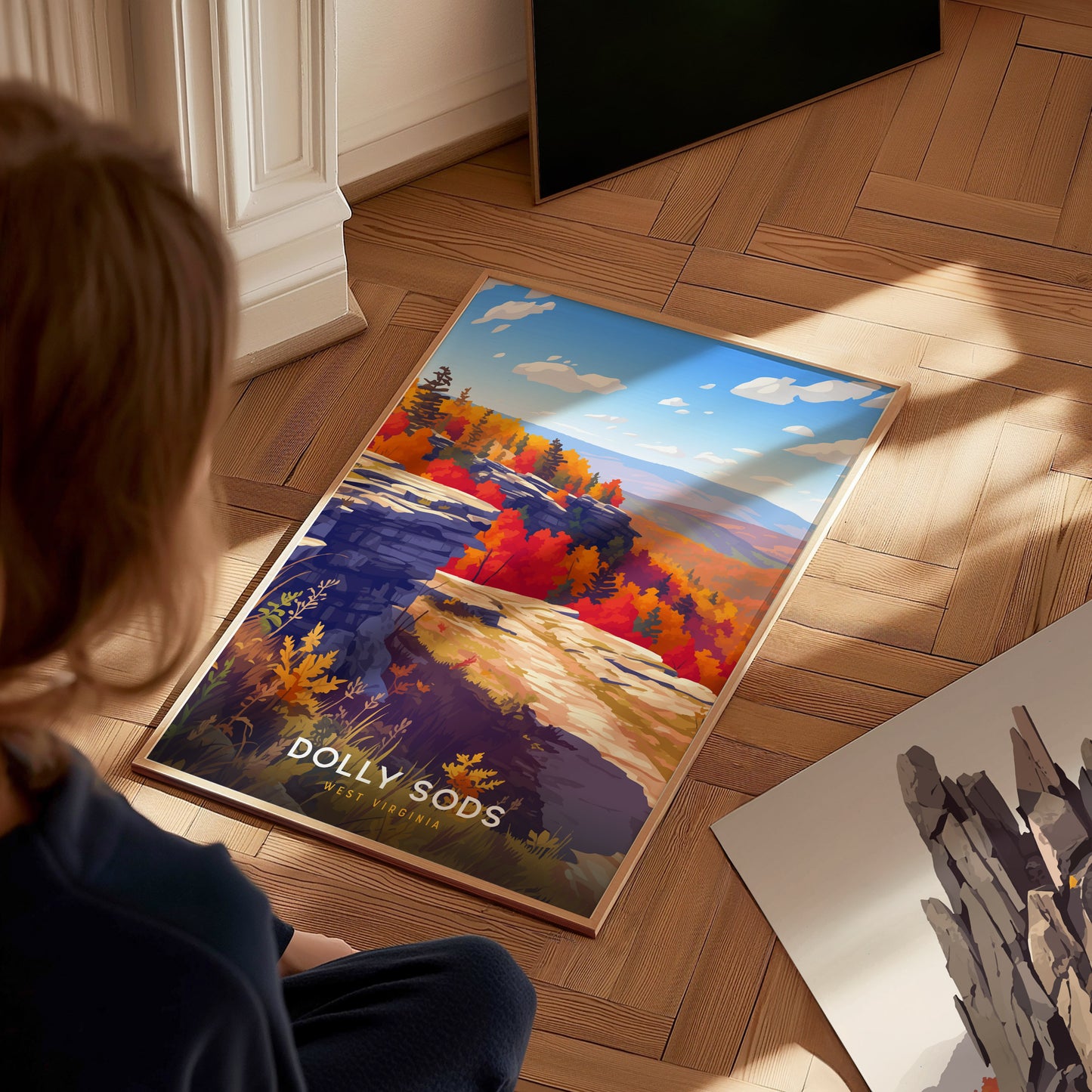 A child sits on a wooden floor, gazing at a vibrant poster of Dolly Sods, West Virginia. The artwork features colorful autumn foliage, rocky cliffs, and a clear blue sky, capturing the beauty of the natural landscape.