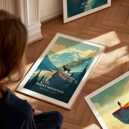 A person looks at a framed Rocky Mountain National Park poster on a wooden floor, depicting an elk on a cliff with mountains and trees in the background.