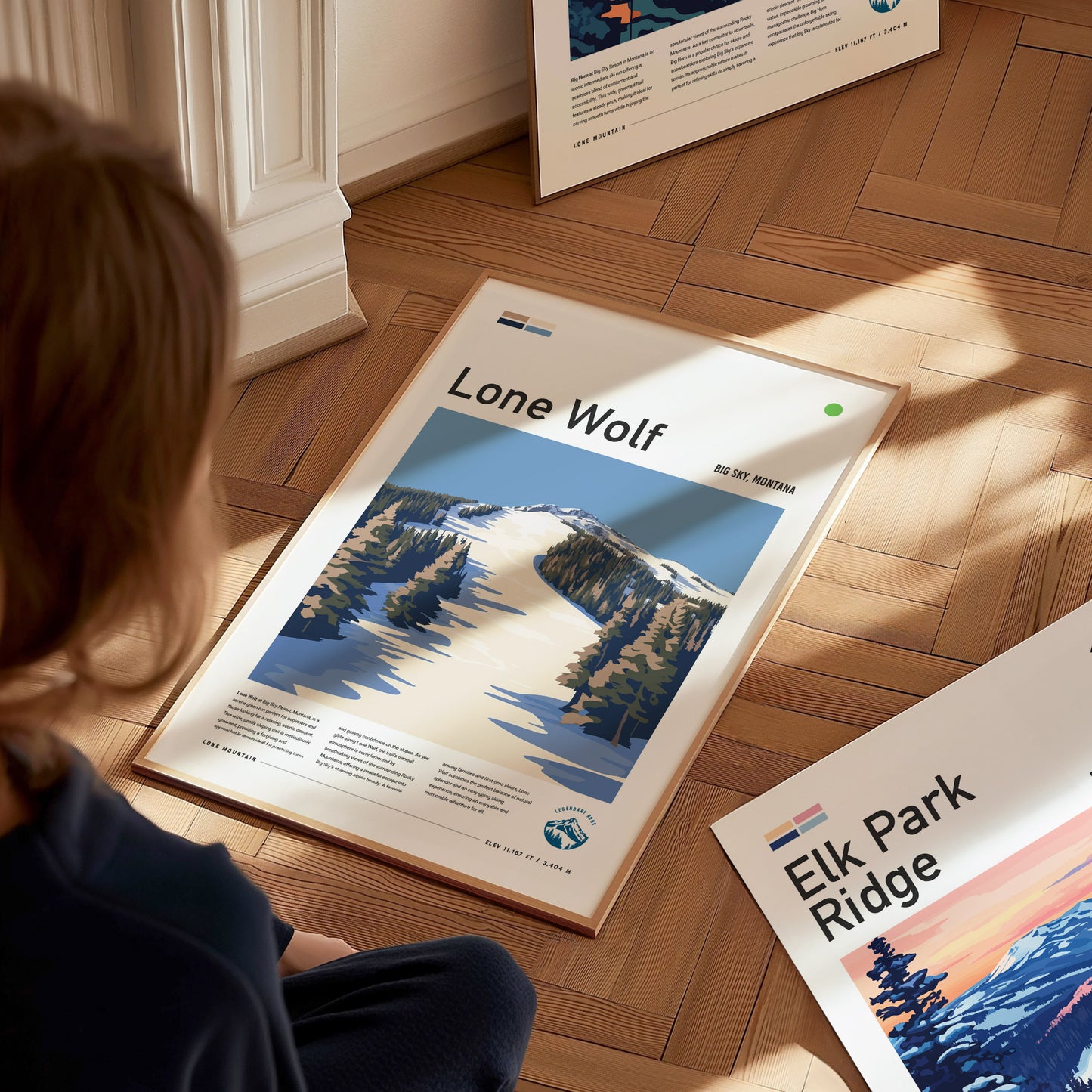 A person sits on a wooden floor, looking at a framed Lone Wolf poster featuring a snowy mountain scene. Another poster titled Elk Park Ridge is partially visible nearby.