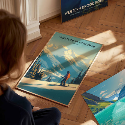 A person sits on a wooden floor, looking at a Whistler Blackcomb poster featuring a skier against snow-covered mountains. Sunlight streams in, illuminating the vibrant artwork.