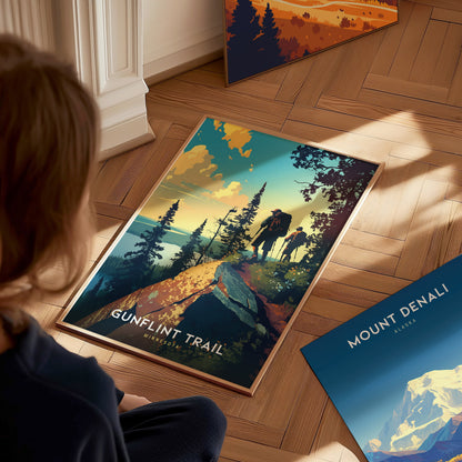 A person observes a Gunflint Trail, Minnesota poster on a wooden floor, featuring hikers on a scenic forested cliff. Nearby, a Mount Denali, Alaska poster is partially visible.