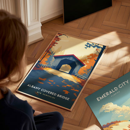 A child sits on a wooden floor, looking at a colorful poster of the Albany Covered Bridge in New Hampshire, featuring vibrant autumn foliage in shades of orange, red, and gold.