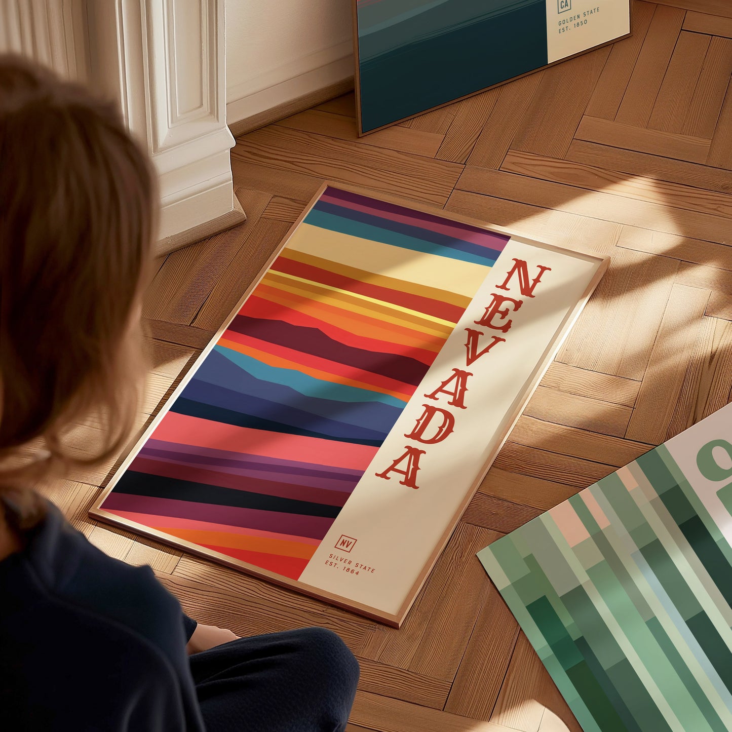 A person sits on a wooden floor, looking at a colorful Nevada poster featuring bold, horizontal stripes and the word Nevada in red. Other posters are partially visible nearby.