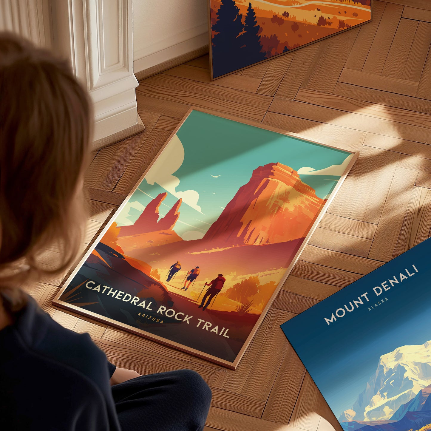 A person sits on a wooden floor, looking at a colorful Cathedral Rock Trail poster depicting hikers and red cliffs. Nearby, another poster shows Mount Denali, Alaska.