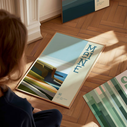 A person observes a minimalist poster of Maine on a wooden floor, featuring abstract coastal imagery and bold lettering. Sunlight casts soft shadows across the scene.