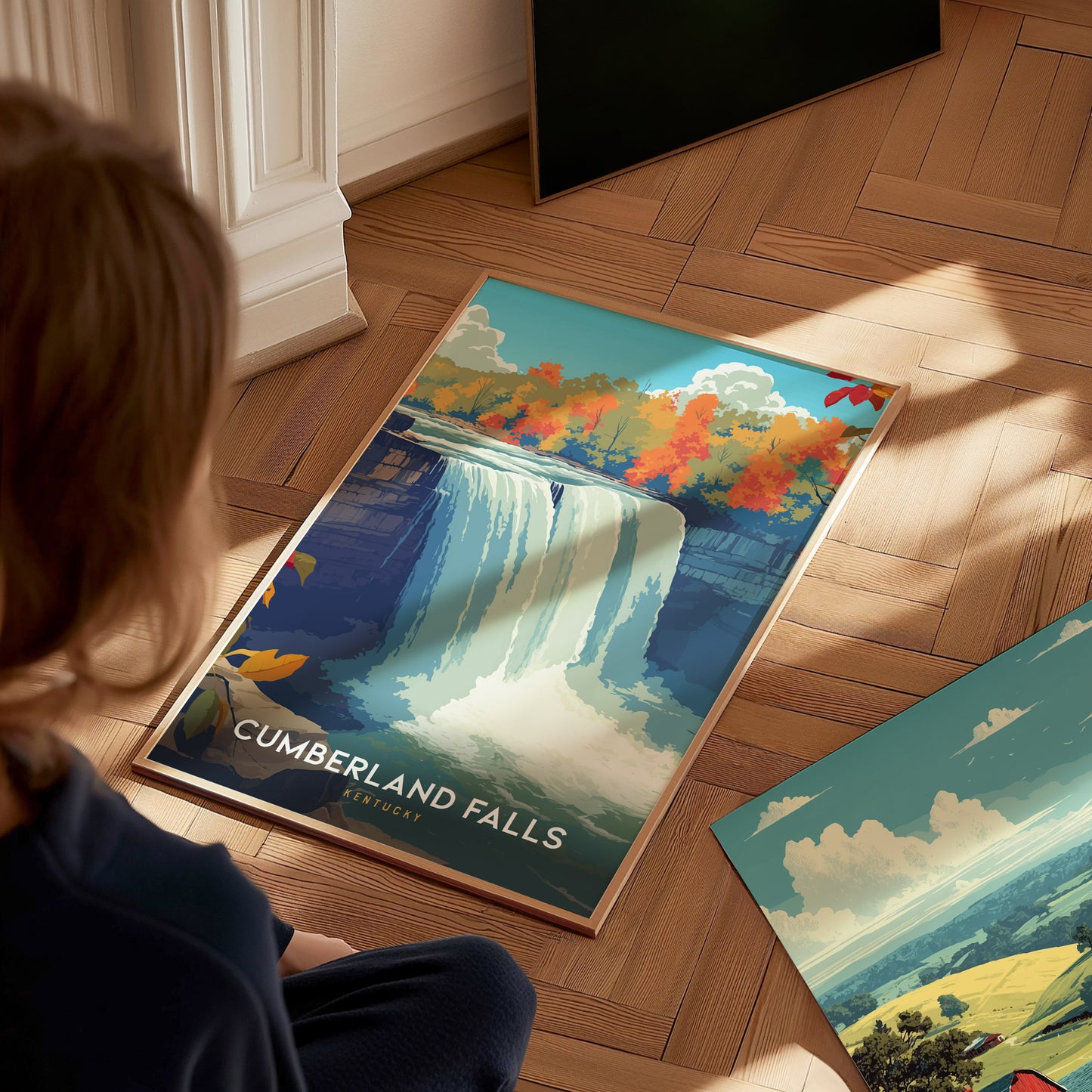A child sits on a wooden floor, gazing at a colorful poster of Cumberland Falls, featuring a majestic waterfall surrounded by vibrant autumn foliage.