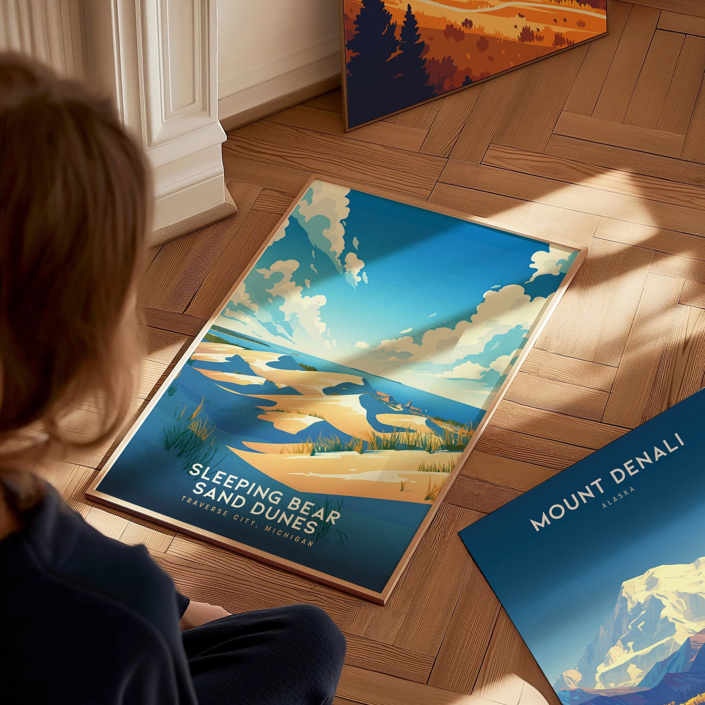 A person looks at a colorful Sleeping Bear Sand Dunes poster on a wooden floor, alongside a Mount Denali print. The room is warmly lit.