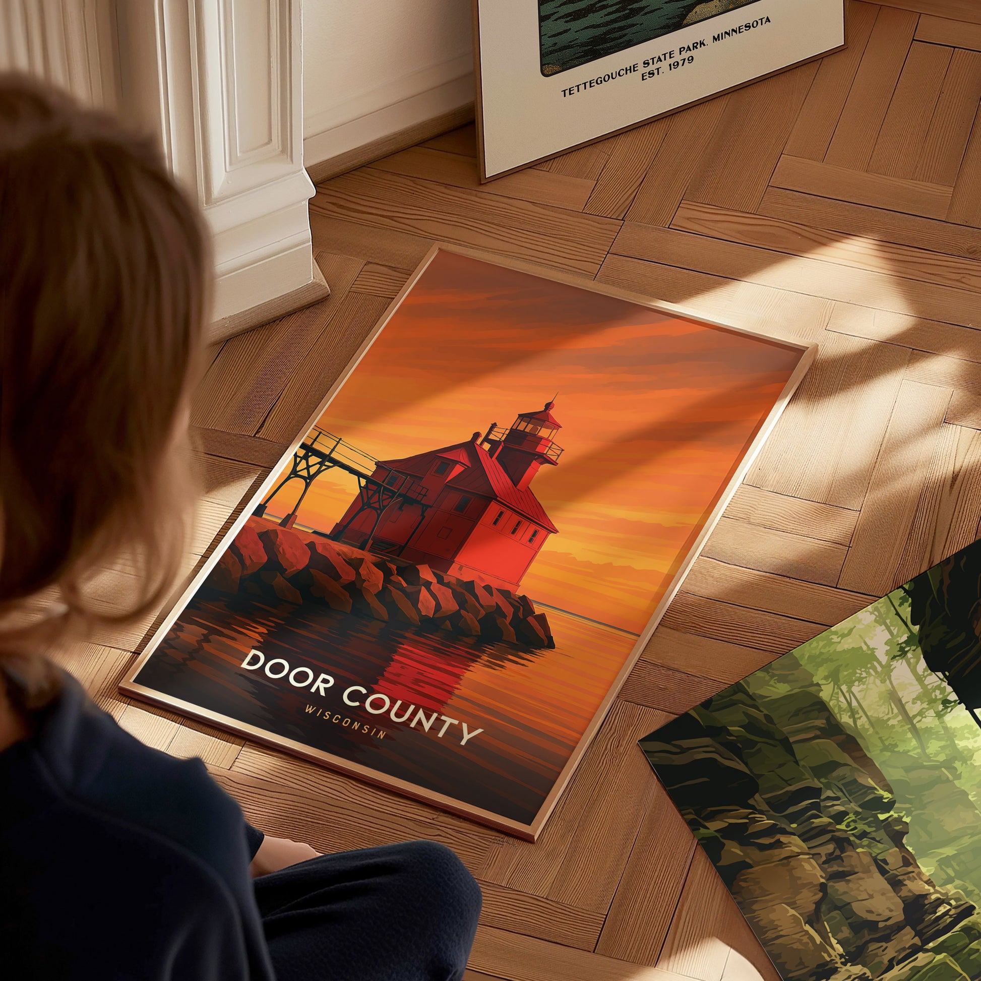 A child sits on a wooden floor, gazing at a framed travel print of Door County, featuring a striking red lighthouse against a vibrant sunset. The artwork showcases warm colors and bold graphic lines, adding a cozy touch to the room.