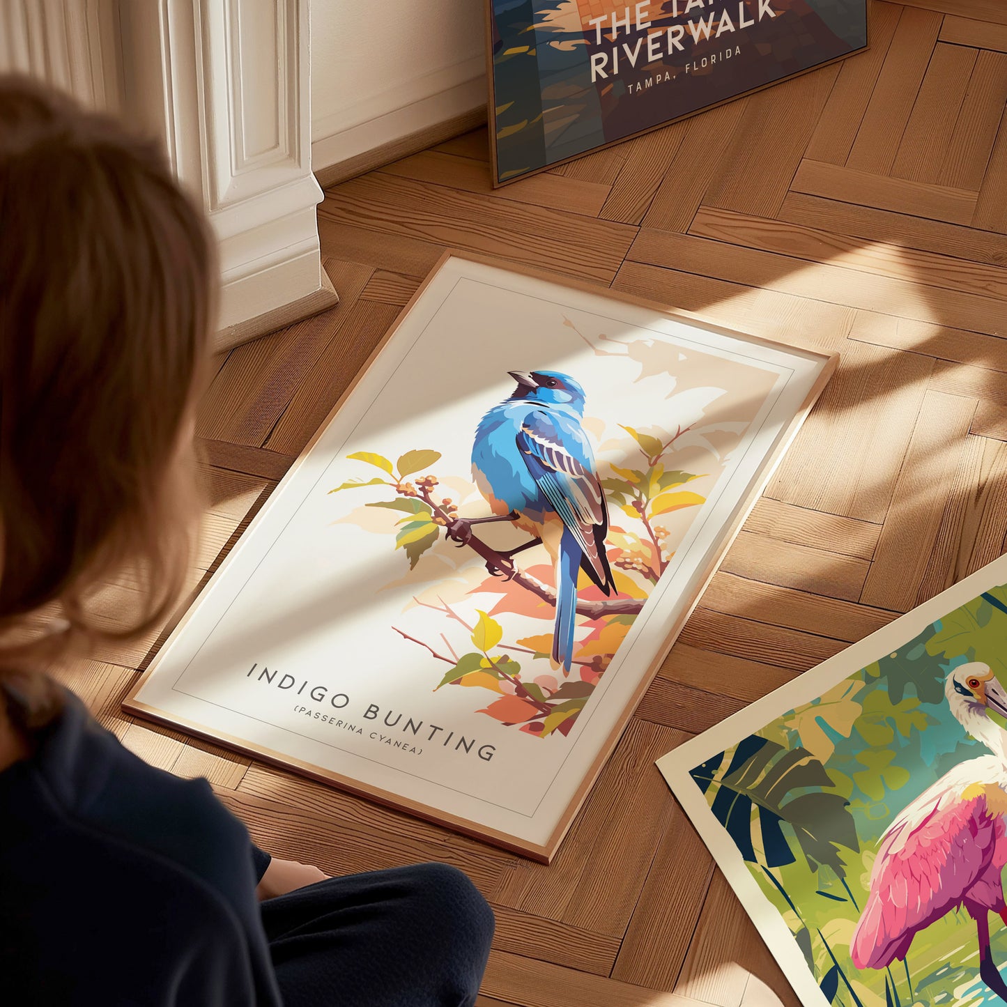 A child sits on a wooden floor, admiring a framed art print featuring a vibrant blue indigo bunting perched on branches with autumn leaves. Other colorful art prints are spread out nearby, adding to the creative atmosphere.