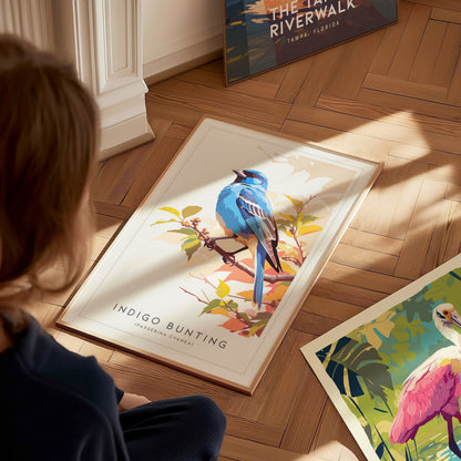 A child sits on a wooden floor, admiring a framed art print featuring a vibrant blue indigo bunting perched on branches with autumn leaves. Other colorful art prints are spread out nearby, adding to the creative atmosphere.