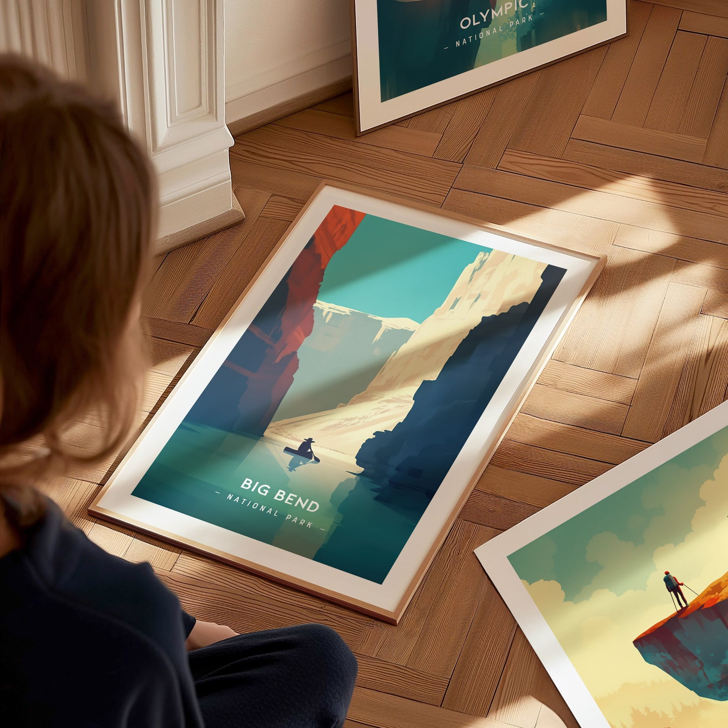A person sits on the floor, admiring a framed Big Bend National Park poster featuring a minimalist landscape with canyons and a figure on a river.