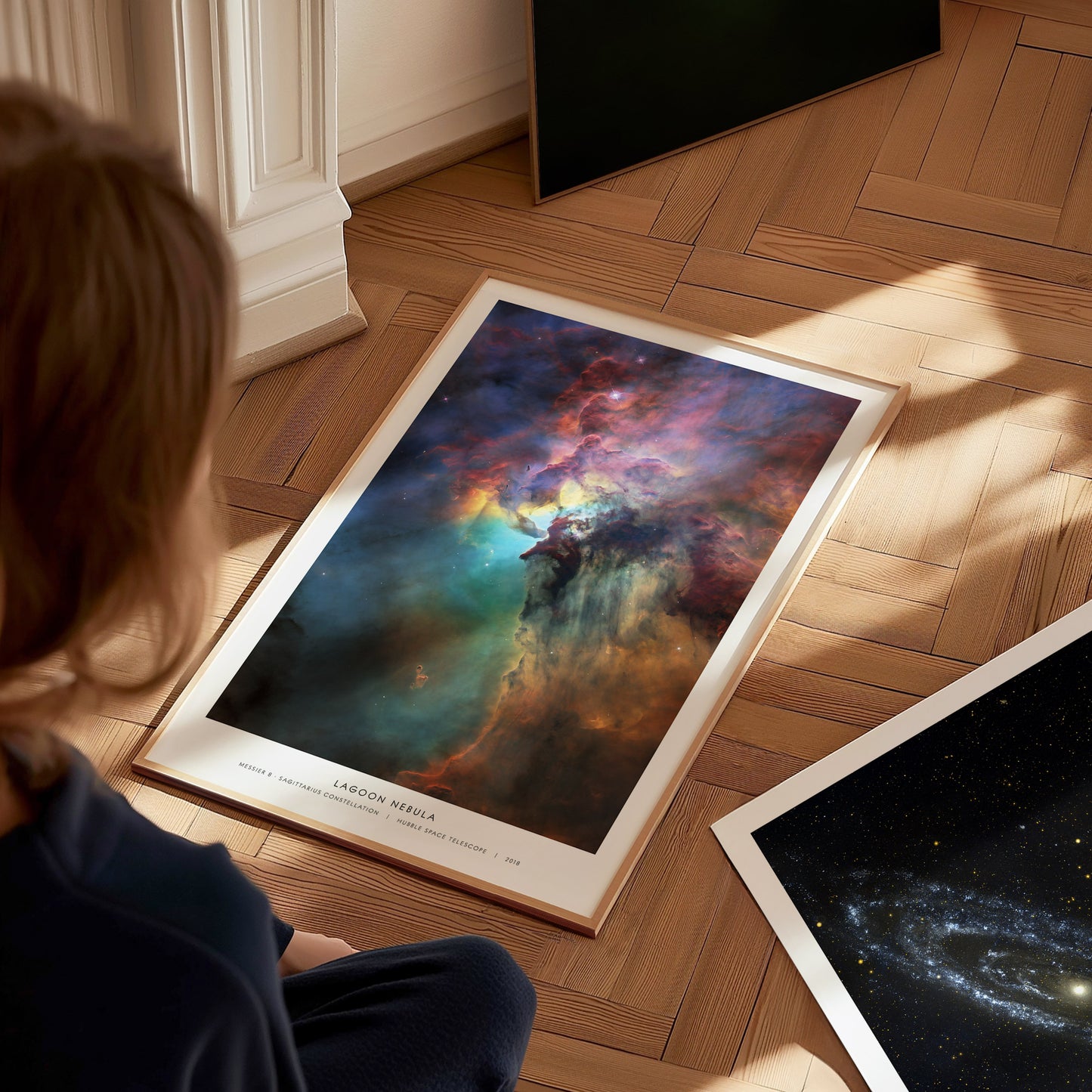A child sits on a wooden floor, gazing at a large, vibrant poster featuring a colorful depiction of the Lagoon Nebula, with swirling clouds of blues, purples, and oranges.
