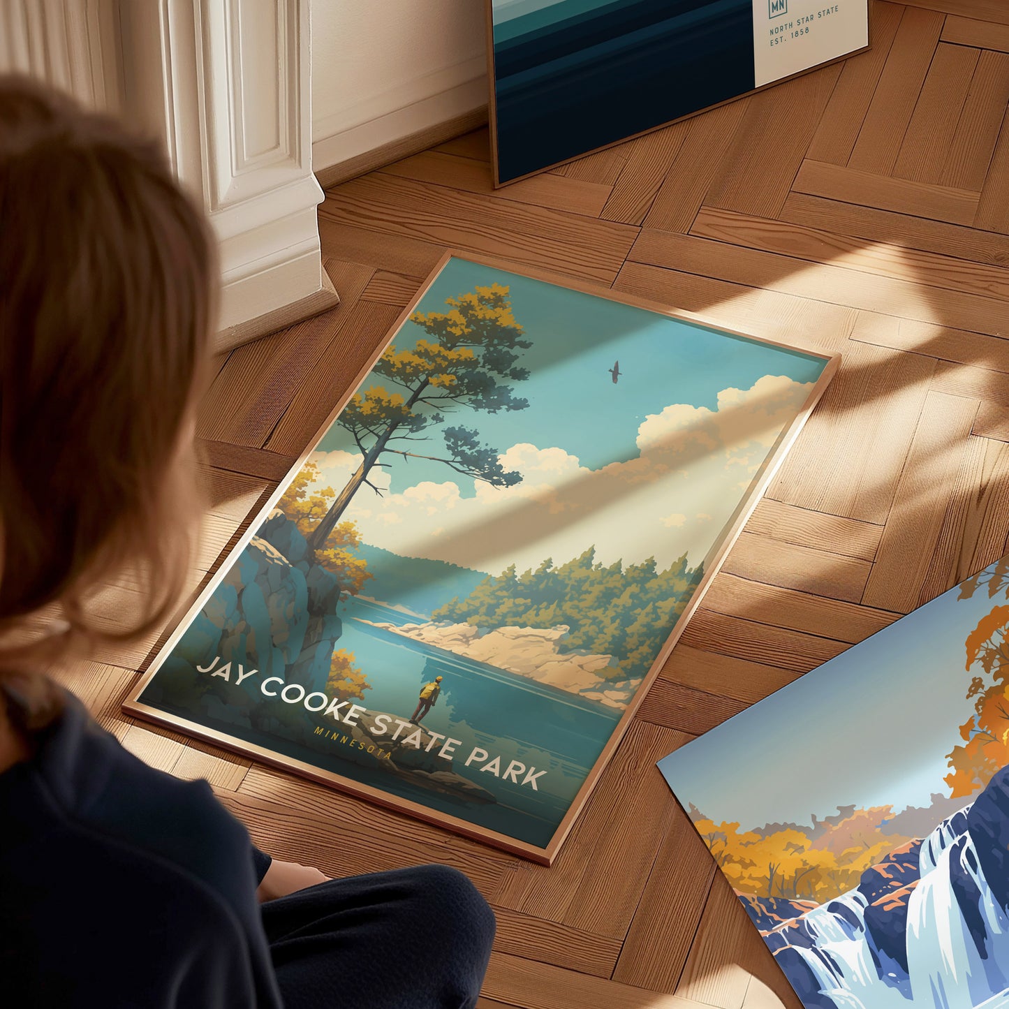 A person observes a Jay Cooke State Park poster on a wooden floor, depicting a scenic landscape with trees, a river, and a bird in the sky.