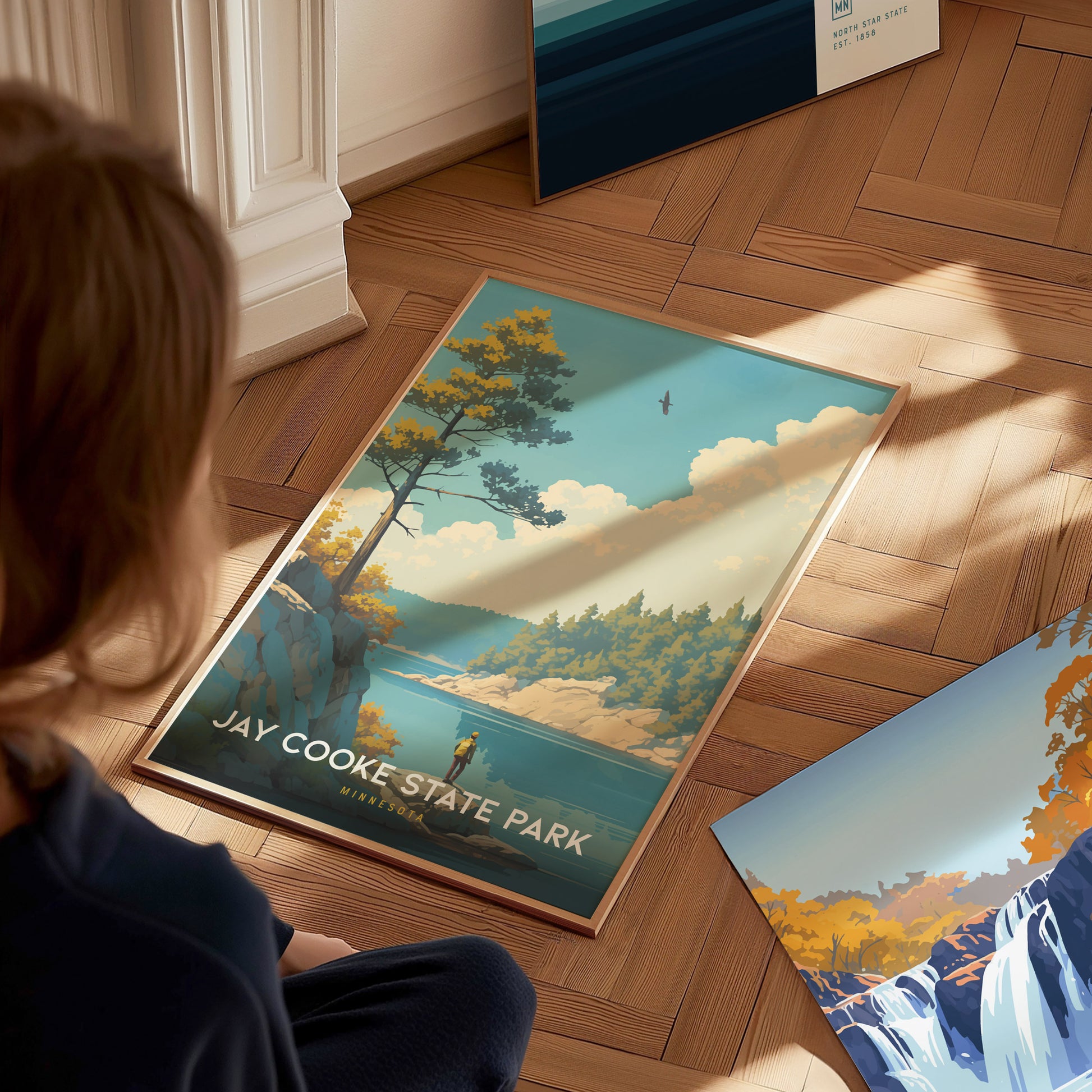 A person observes a Jay Cooke State Park poster on a wooden floor, depicting a scenic landscape with trees, a river, and a bird in the sky.