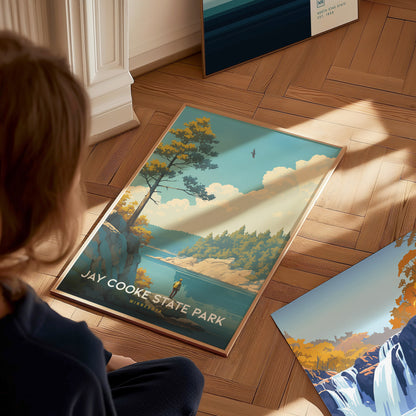 A person observes a Jay Cooke State Park poster on a wooden floor, depicting a scenic landscape with trees, a river, and a bird in the sky.