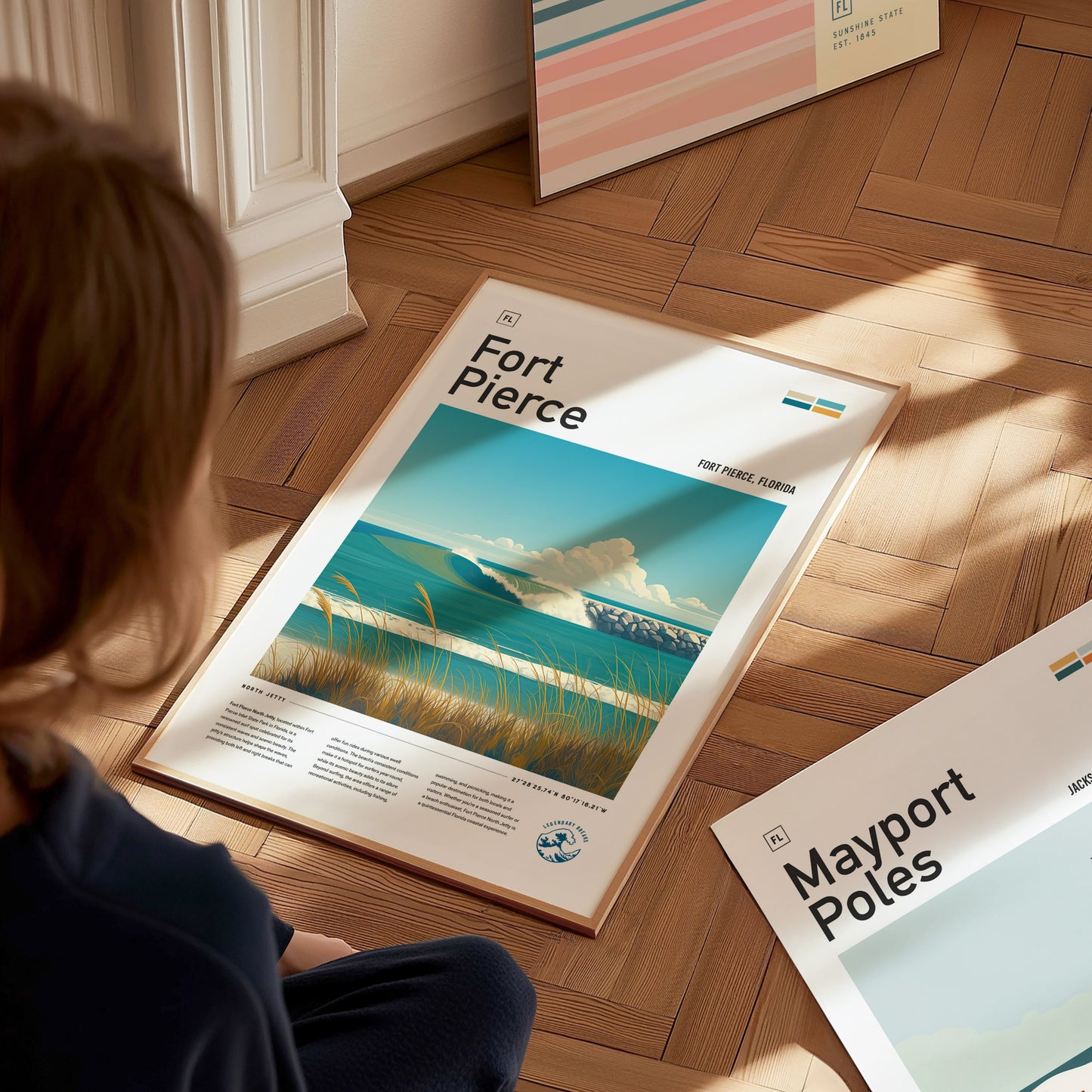 A person sits on a wooden floor gazing at a Fort Pierce poster featuring a coastal scene with waves and clouds. Additional posters are partially visible nearby.