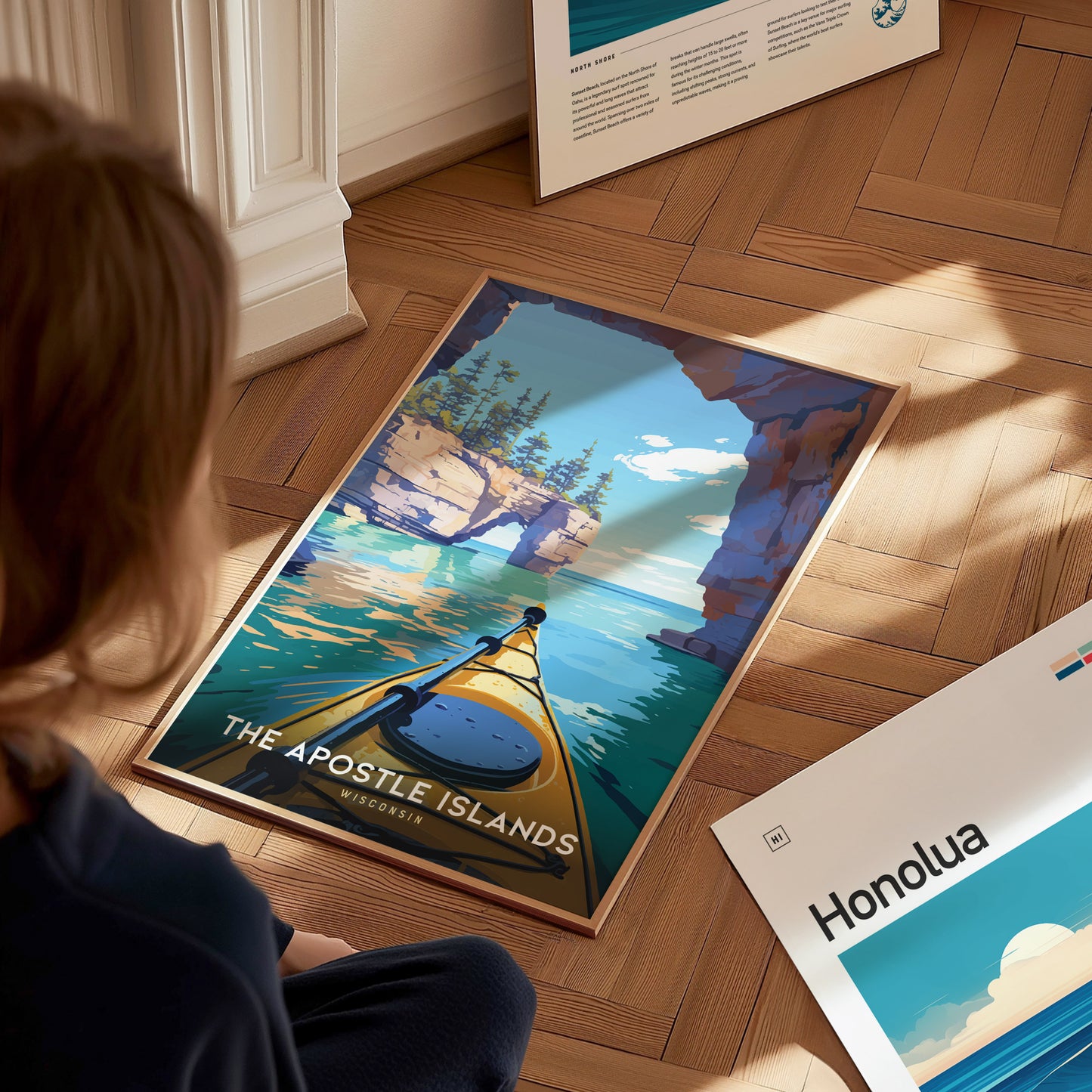 A person sits on a wooden floor, gazing at a vibrant poster depicting a kayak navigating through the Apostle Islands, showcasing cliffs and clear blue waters.