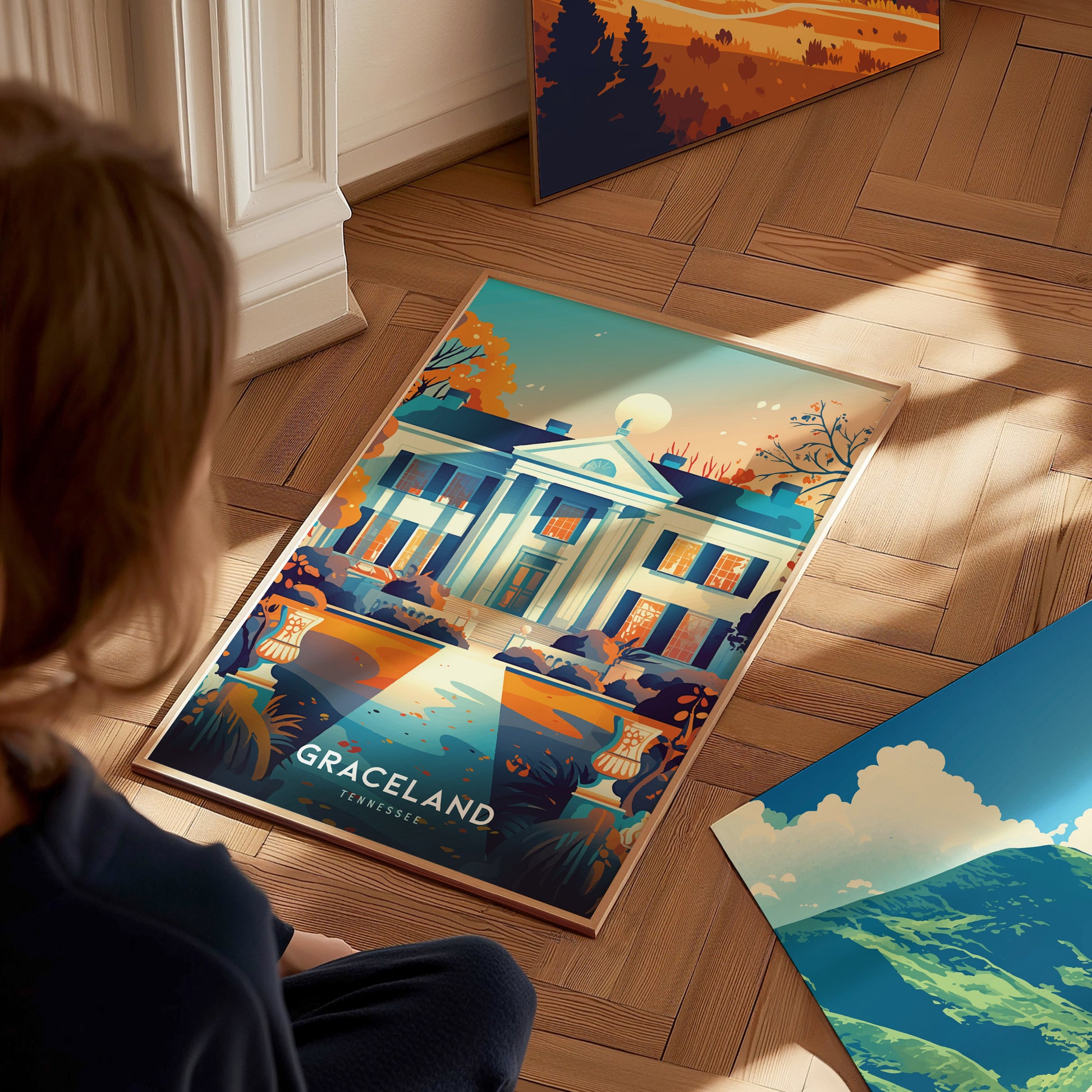 A person sits on a wooden floor, looking at a colorful poster of Graceland, Tennessee. The poster features a vibrant depiction of the iconic mansion amidst autumnal foliage.