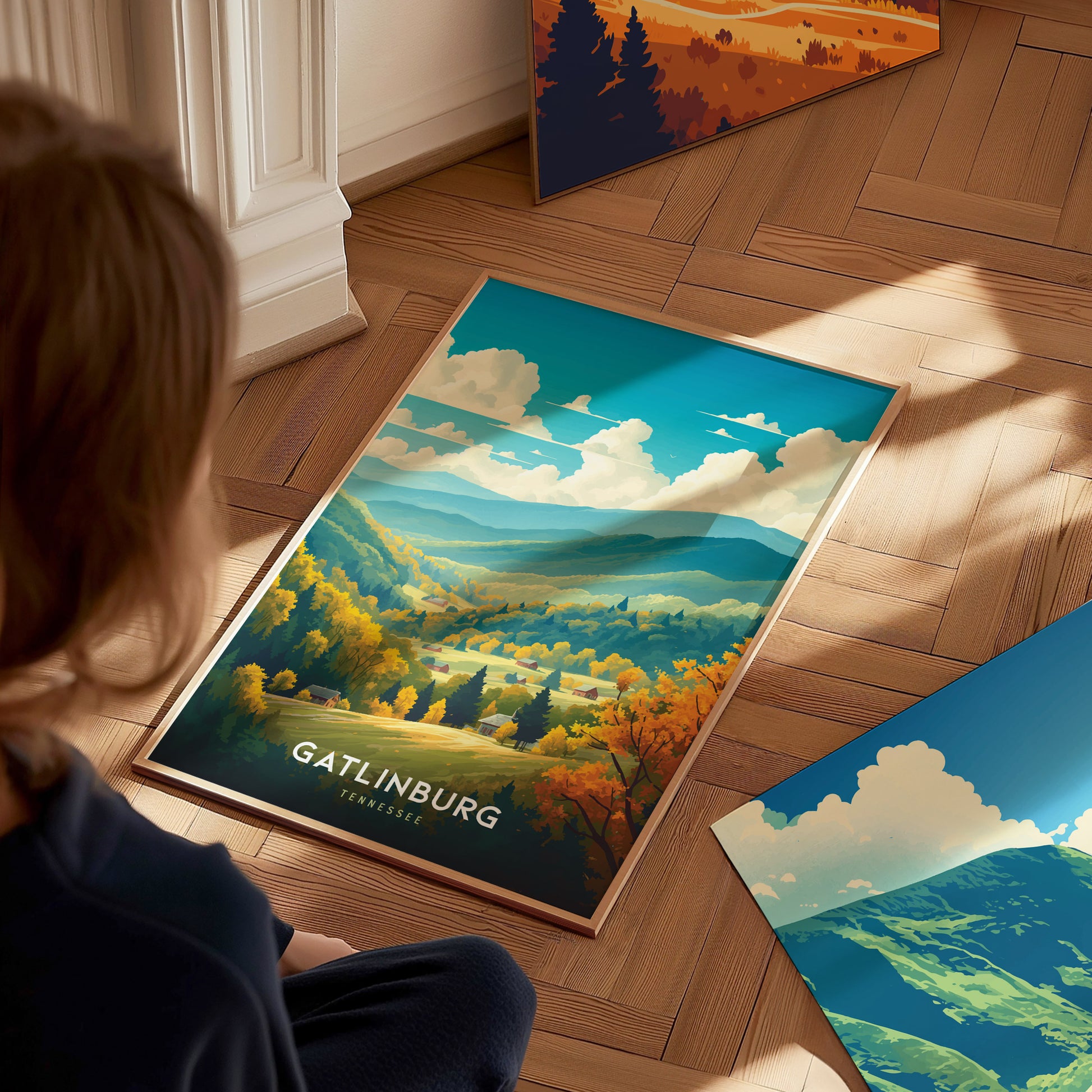 A person observes a colorful Gatlinburg, Tennessee poster featuring lush forests and rolling hills, lying on a wooden floor with sunlight streaming in. Other landscape posters are nearby.