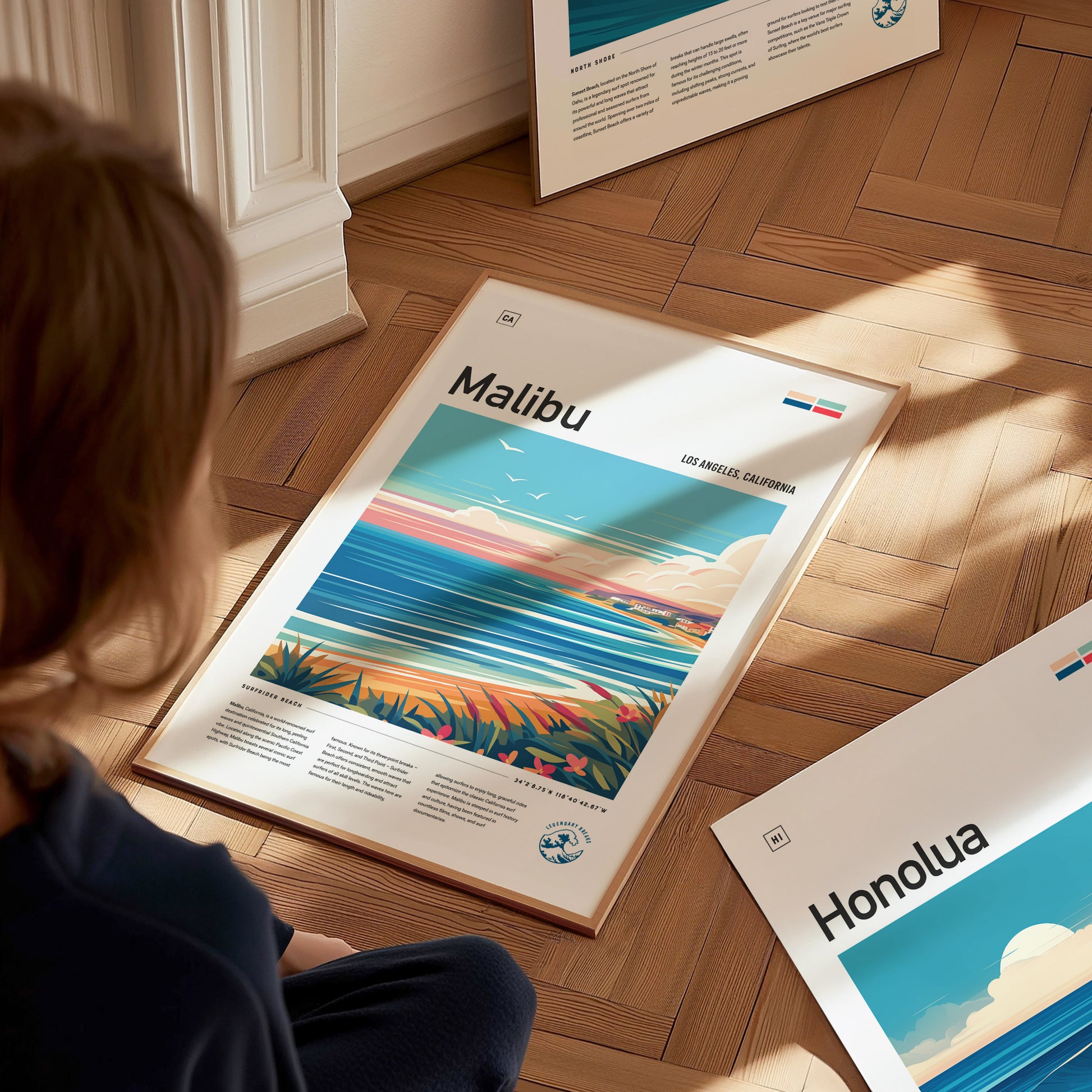 A person looks at a framed poster on the floor titled Malibu, featuring an abstract coastline with vibrant colors and a clear sky.