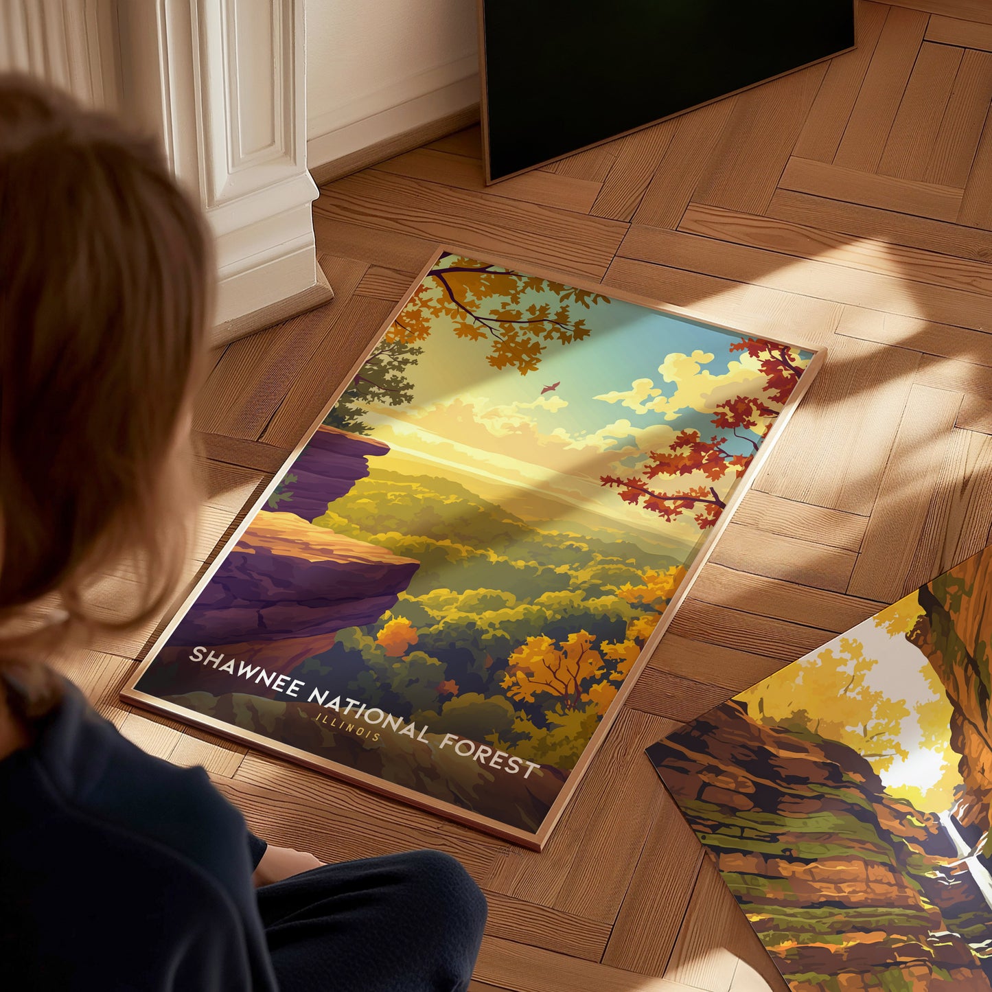 A young person sits on a wooden floor, gazing at a vibrant travel poster of Shawnee National Forest. The artwork showcases a picturesque landscape with colorful autumn foliage, rocky cliffs, and a serene valley bathed in warm sunlight.