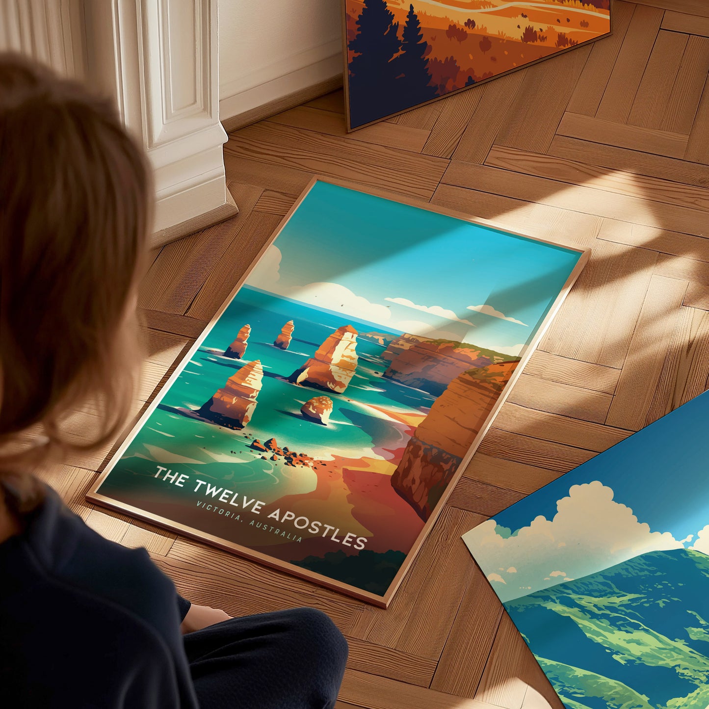 A person sits on a wooden floor, looking at a vibrant poster of the Twelve Apostles with turquoise waters and limestone stacks. Other colorful artwork is partially visible nearby.