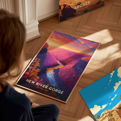 A person sits on a wooden floor looking at a vibrant New River Gorge National Park poster featuring colorful canyon and river scenery. Other posters are partially visible nearby.