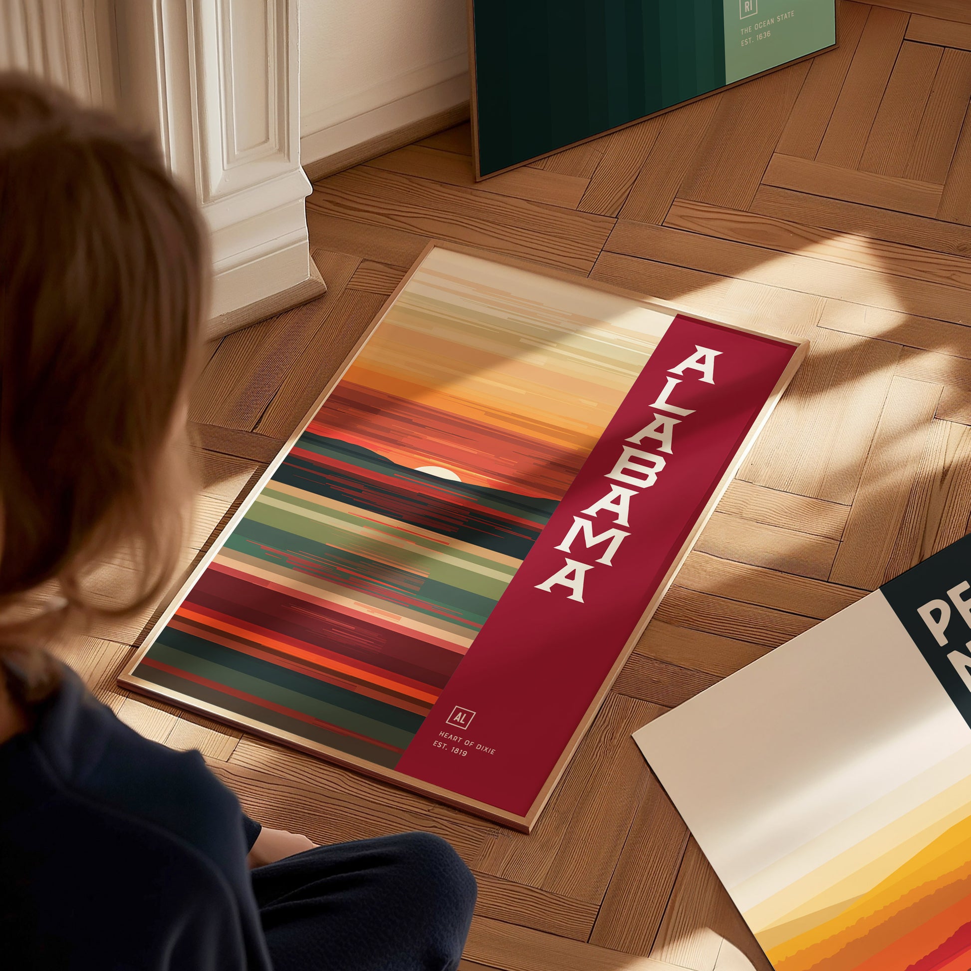 A person sits on a wooden floor, looking at an abstract Alabama poster with vibrant red, orange, and green stripes. The word Alabama is prominently displayed in bold white letters.