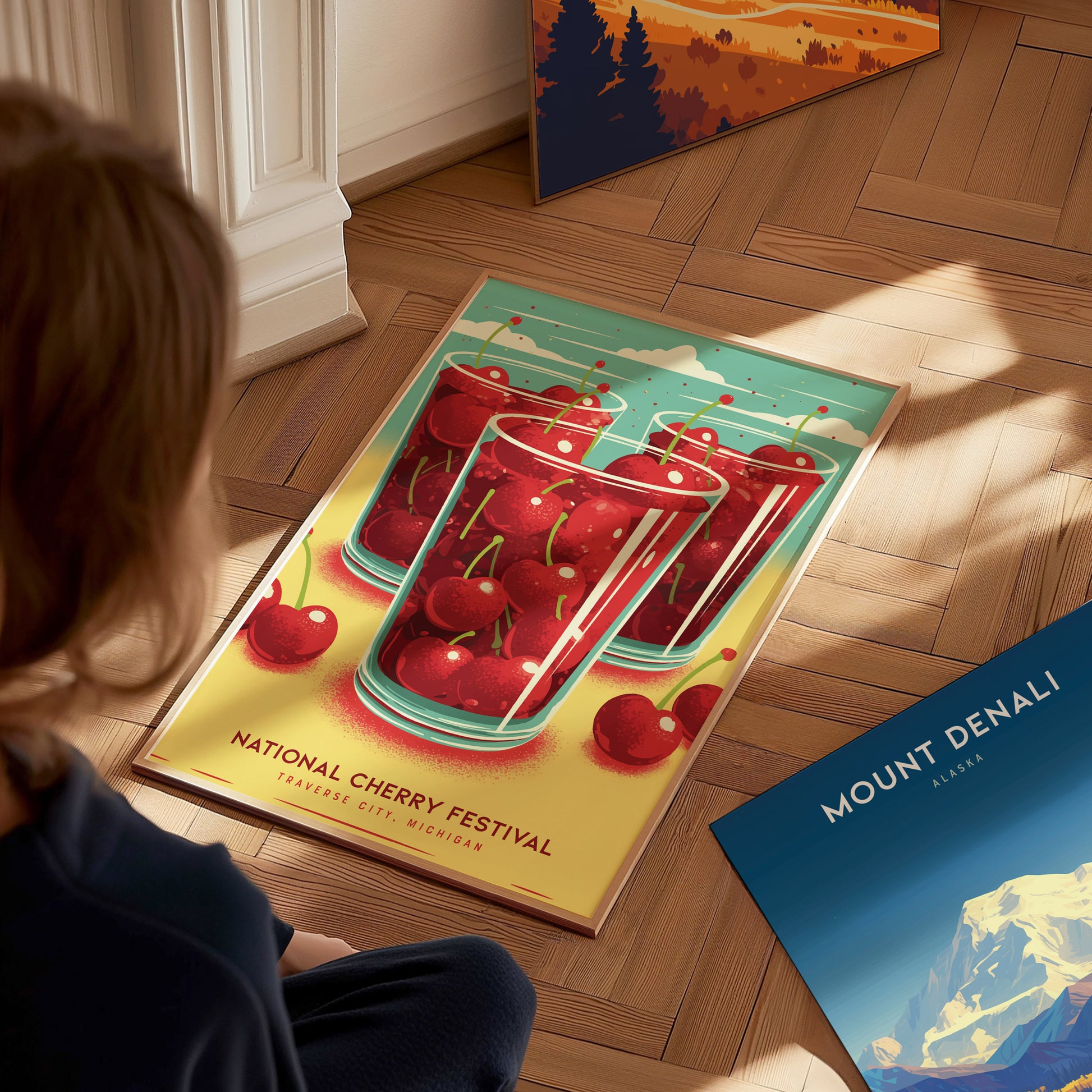 A person observes a colorful National Cherry Festival poster featuring glasses filled with cherries, placed on a wooden floor alongside a Mount Denali print.
