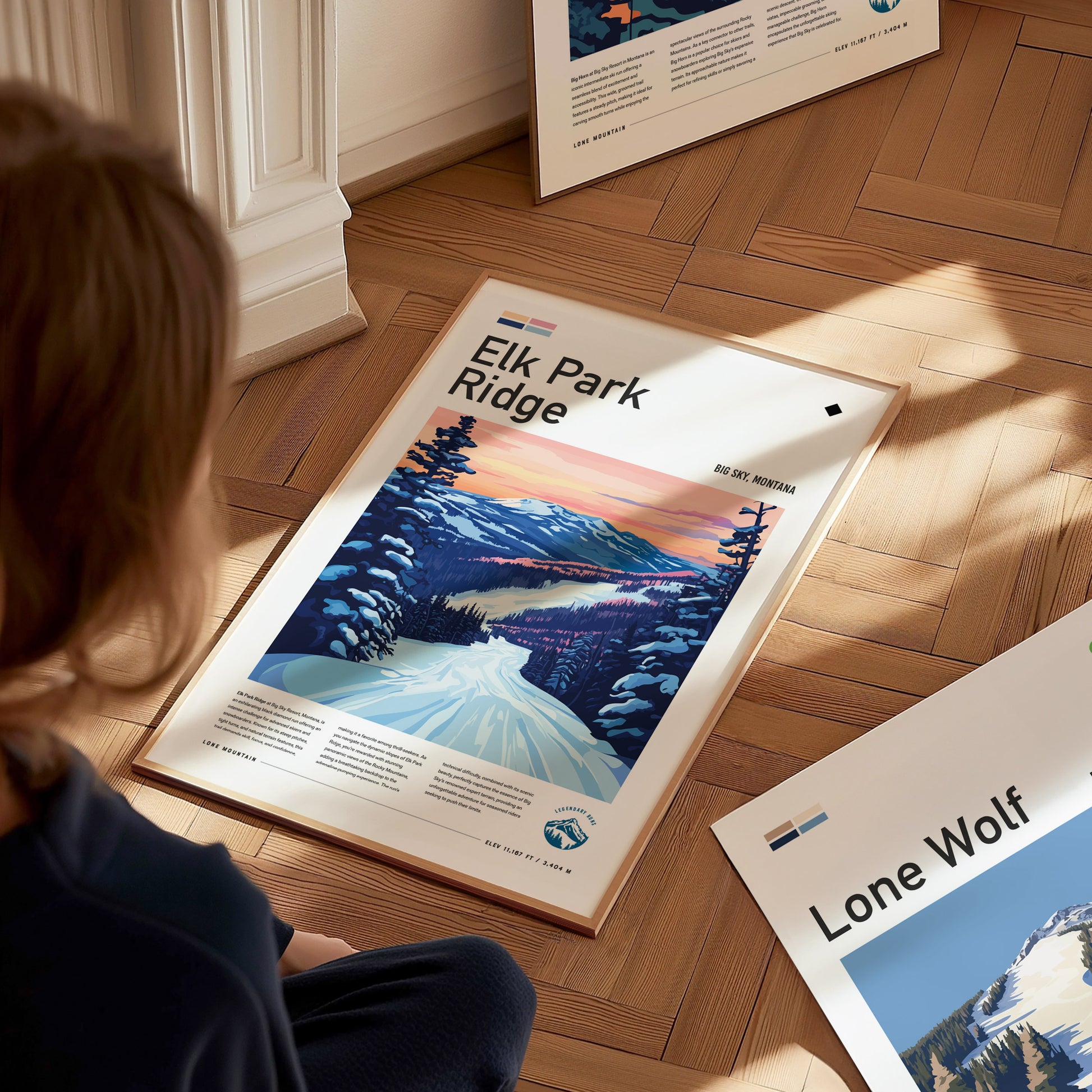 A person sits on a wooden floor viewing a framed Elk Park Ridge ski poster, depicting a snowy mountain landscape with a vibrant sunset. Other posters are scattered around.