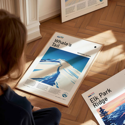 A person sits on a wooden floor observing a framed Whales Tail Breckenridge poster, featuring a stylized snowy mountain scene, with another poster partially visible nearby.