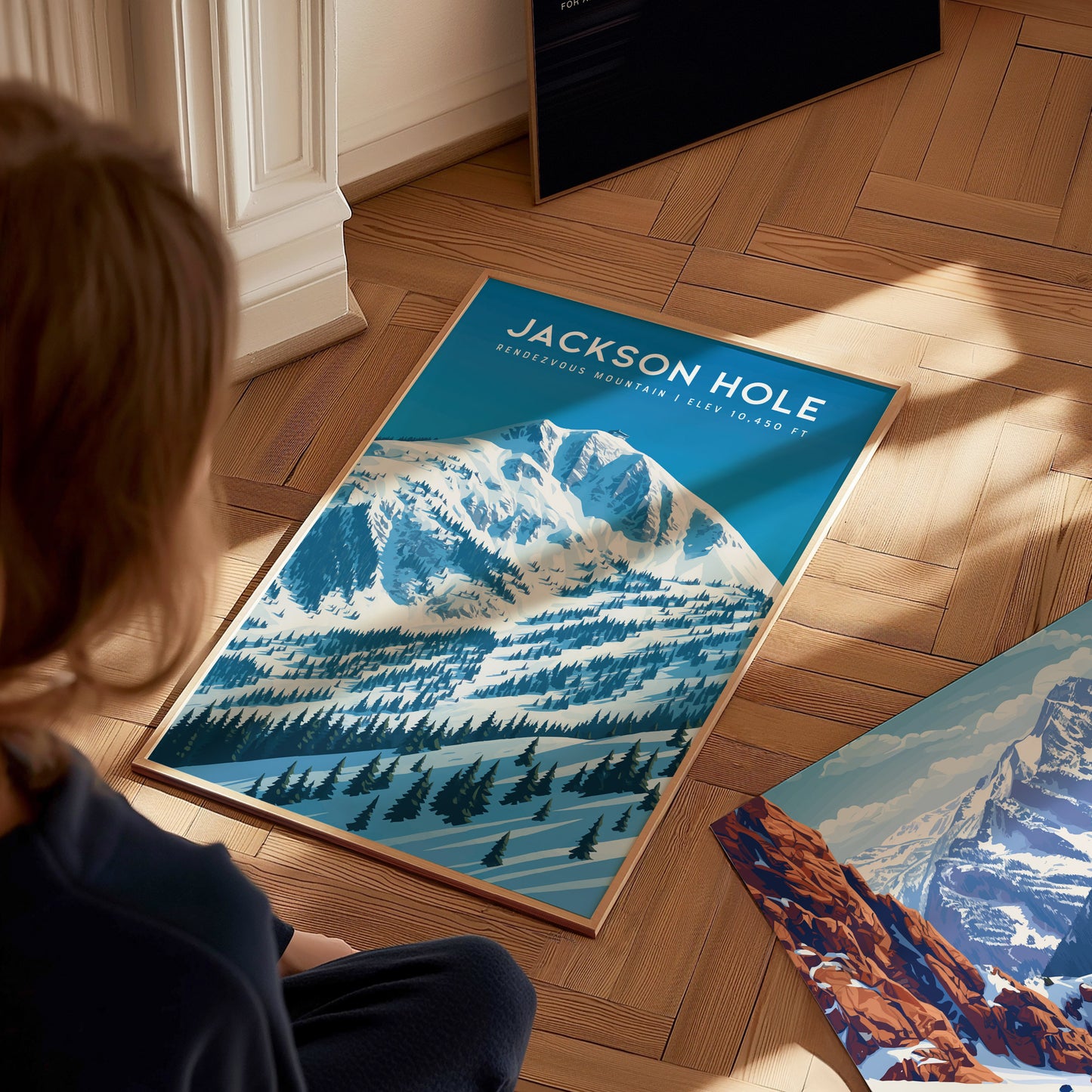 A person sits on a wooden floor, looking at a poster of Jackson Hole featuring snow-capped mountains and pine trees. The vibrant artwork highlights the majestic landscape, with shades of blue and white creating a serene atmosphere.