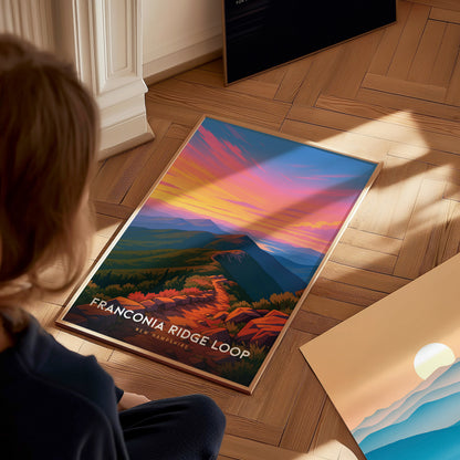A person sits on a wooden floor, gazing at a vibrant poster titled Franconia Ridge Loop, which features a colorful sunset over a mountainous landscape.