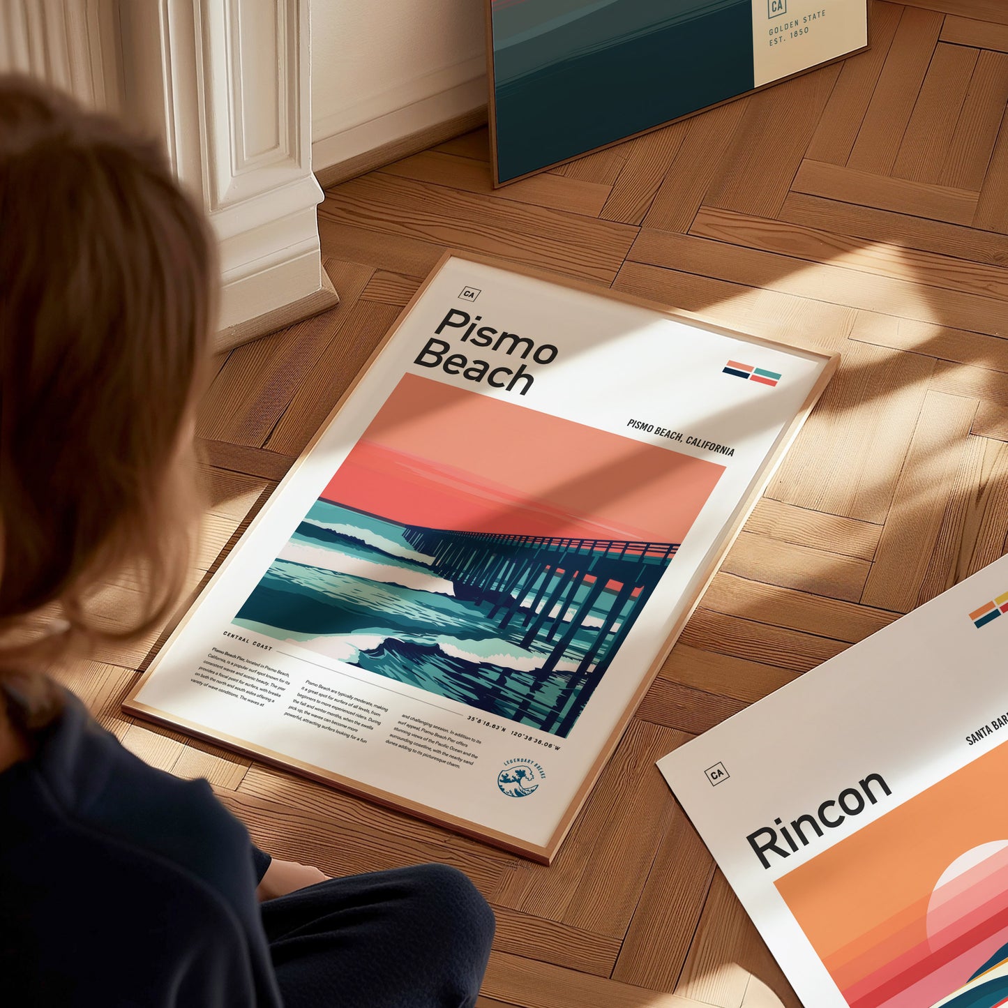A person sits on a wooden floor, looking at a Pismo Beach poster featuring a pier against a vibrant sunset. Another poster titled Rincon is partially visible nearby.