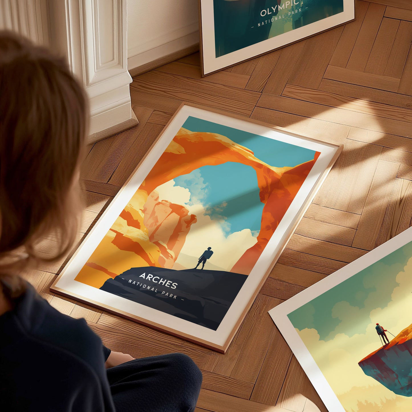 A framed poster of Arches National Park lies on a wooden floor, depicting a lone figure beneath Delicate Arch against a vivid sky. A person observes the poster closely.