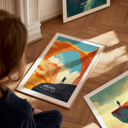A framed poster of Arches National Park lies on a wooden floor, depicting a lone figure beneath Delicate Arch against a vivid sky. A person observes the poster closely.