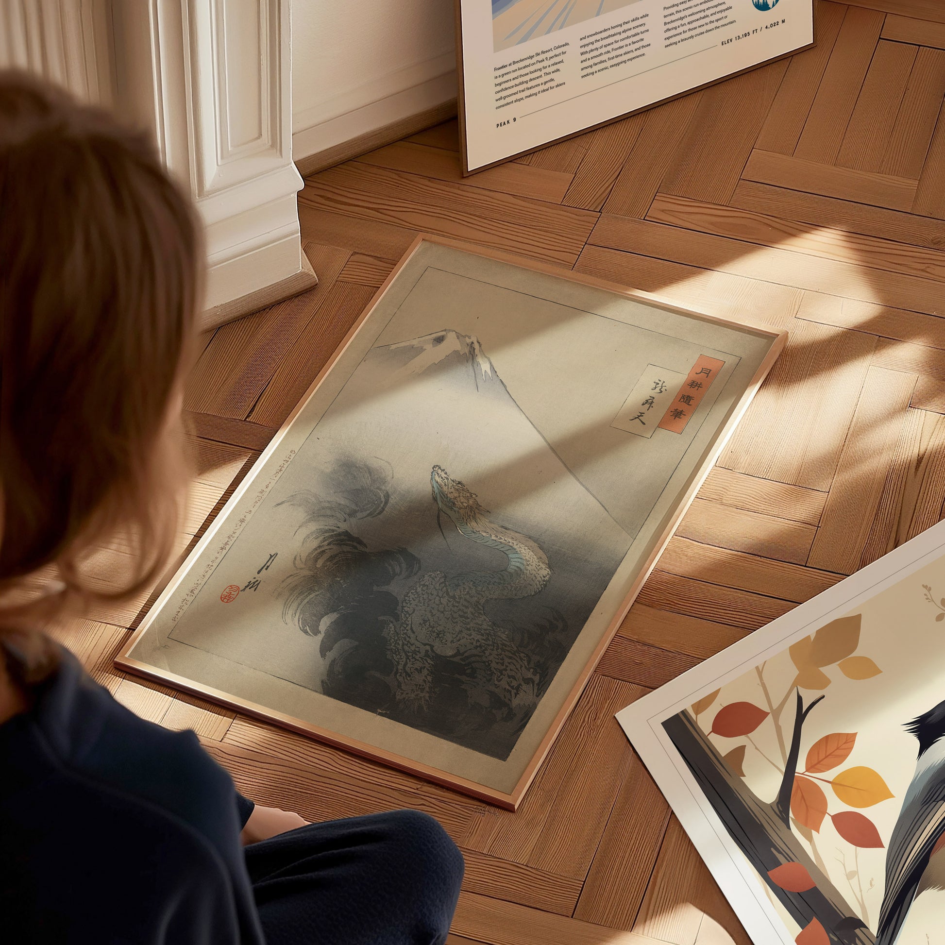 A person sits on a wooden floor, looking at a framed art piece featuring a dragon ascending near Mount Fuji. Another artwork with a bird and leaves is partially visible.