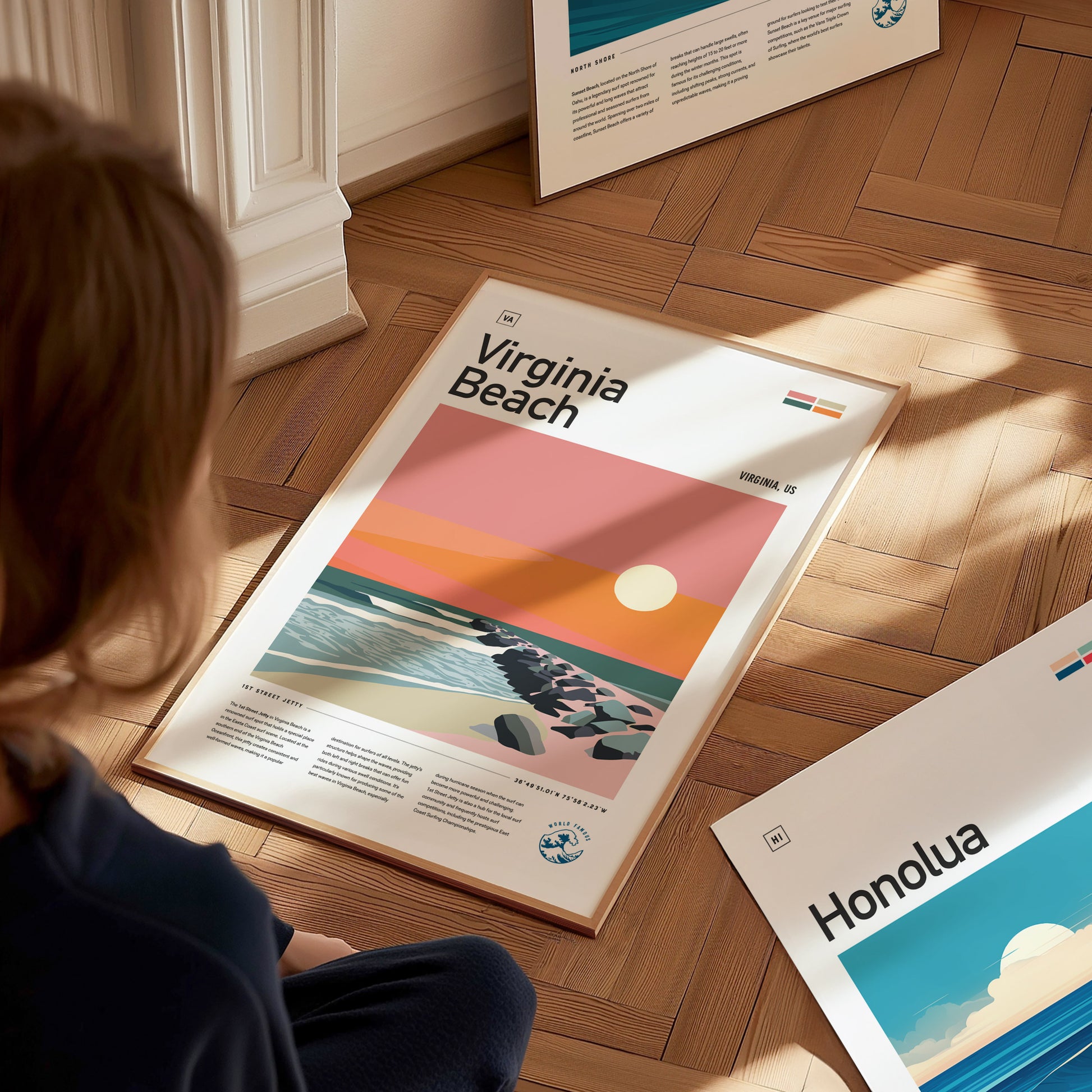 A person observes a vibrant Virginia Beach poster on a wooden floor, featuring a stylized sunset over the ocean and jetty rocks. Other posters are partially visible nearby.