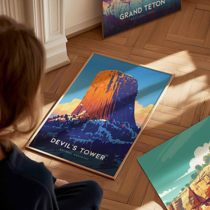 A person looks at a vibrant Devils Tower poster on a wooden floor, with colorful artwork and another poster in the background. Sunlight casts shadows across the scene.