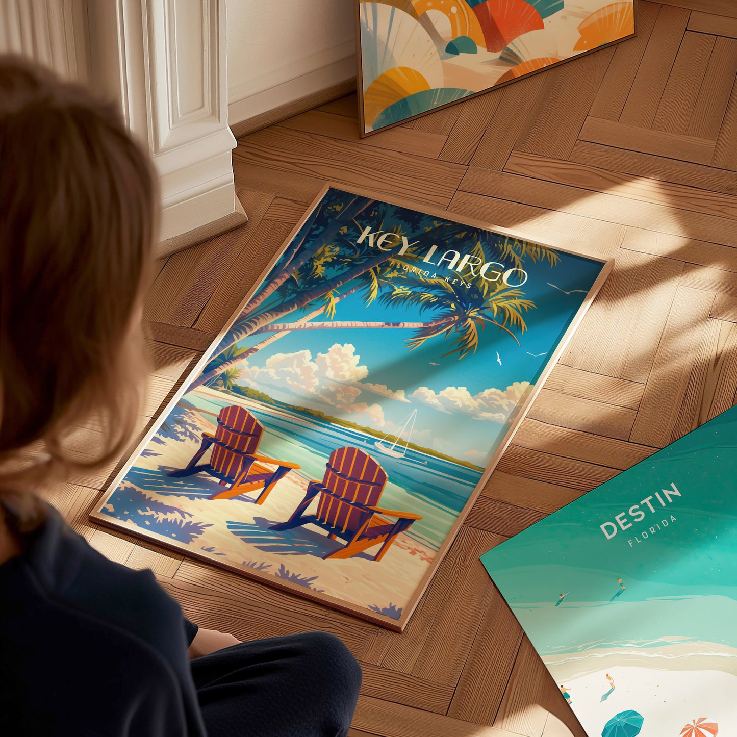 A person looks at a vibrant Key Largo poster featuring two beach chairs under palm trees by the ocean. Nearby, another poster labeled Destin, Florida rests on the wooden floor.