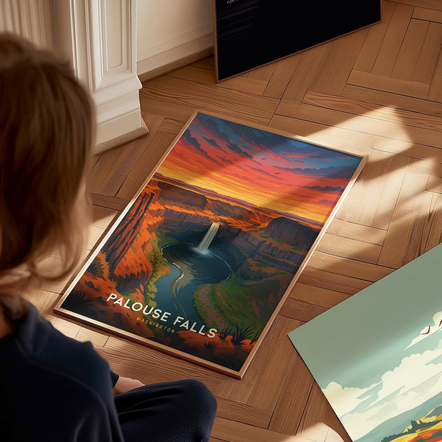 A person sits on a wooden floor, looking at a colorful travel poster of Palouse Falls, featuring a dramatic sunset with vibrant orange and purple hues above a waterfall surrounded by cliffs and lush greenery.