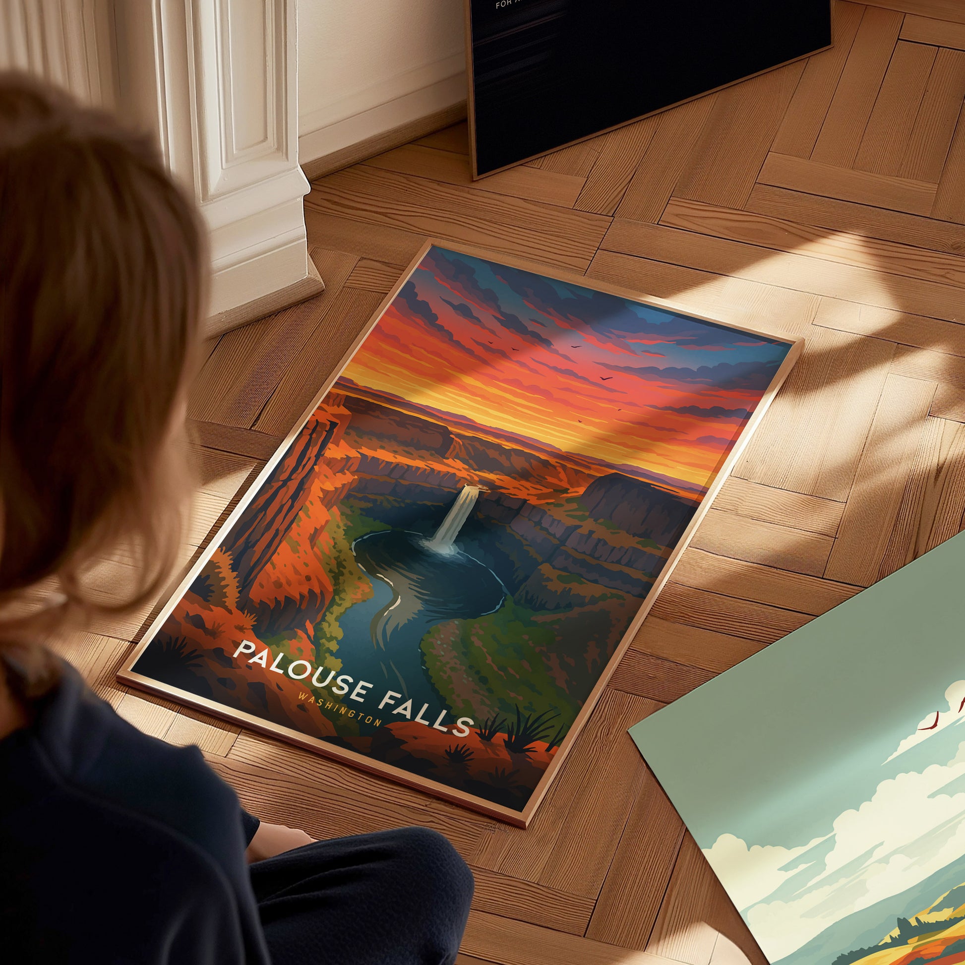 A person sits on a wooden floor, looking at a colorful travel poster of Palouse Falls, featuring a dramatic sunset with vibrant orange and purple hues above a waterfall surrounded by cliffs and lush greenery.