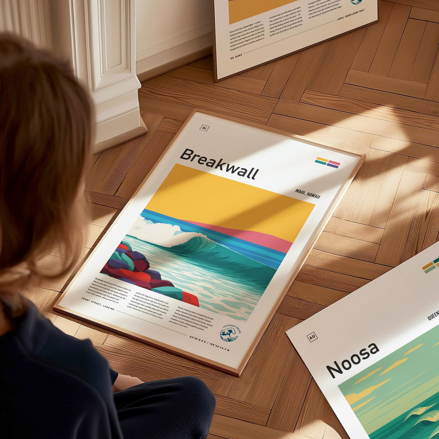 A person sits on a wooden floor, gazing at a framed surf poster titled Breakwall. The artwork depicts a vibrant wave with colorful landforms under a bright yellow sky, highlighting a lively surf culture aesthetic.