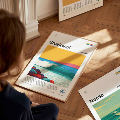 A person sits on a wooden floor, gazing at a framed surf poster titled Breakwall. The artwork depicts a vibrant wave with colorful landforms under a bright yellow sky, highlighting a lively surf culture aesthetic.