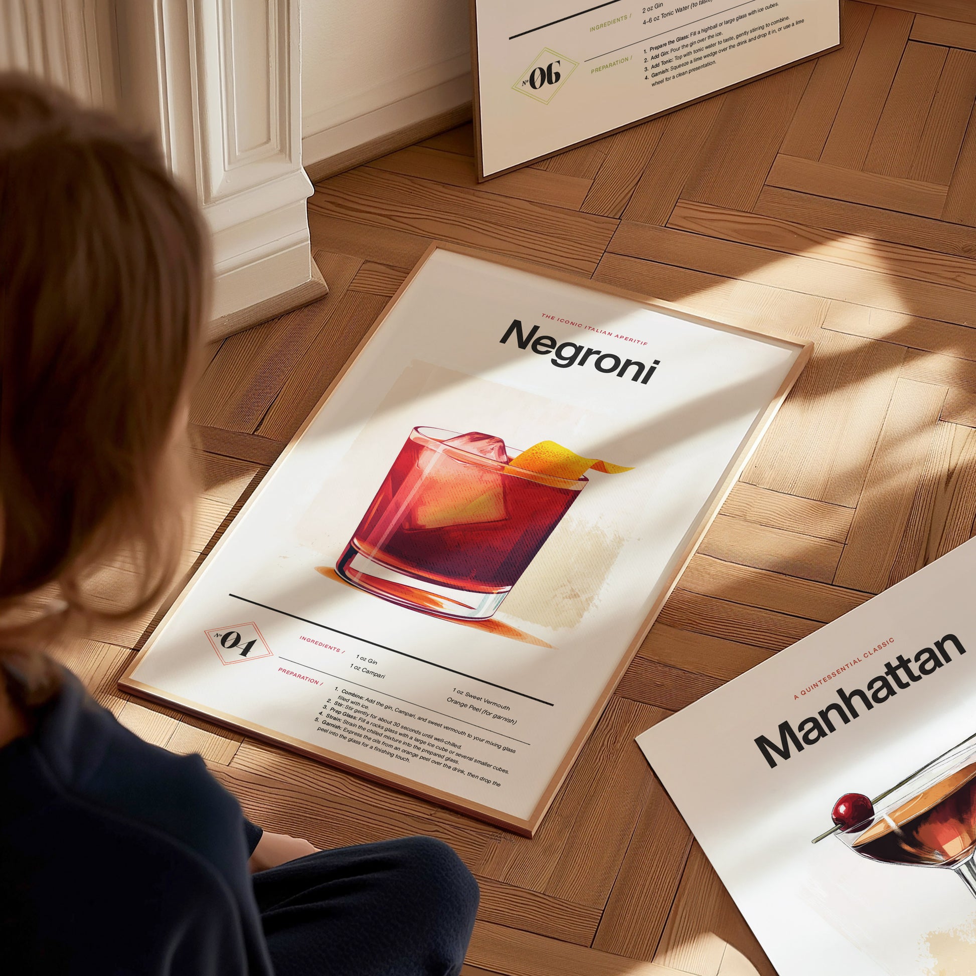 A person observes a Negroni cocktail poster on a wooden floor, featuring a vibrant drink with an orange peel garnish. Another poster titled Manhattan is partially visible nearby.