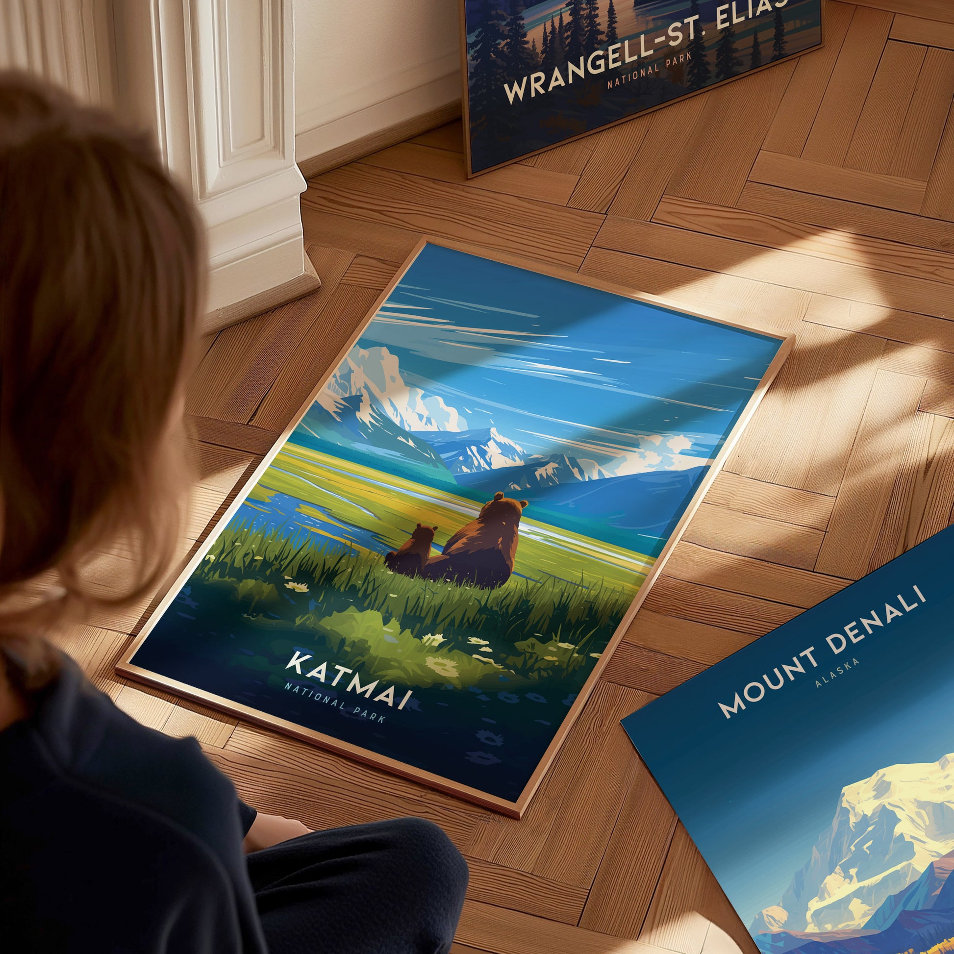 A person sits on a wooden floor, admiring a vibrant Katmai National Park poster featuring a bear and cub against a mountain backdrop. Nearby, Mount Denali and Wrangell-St.