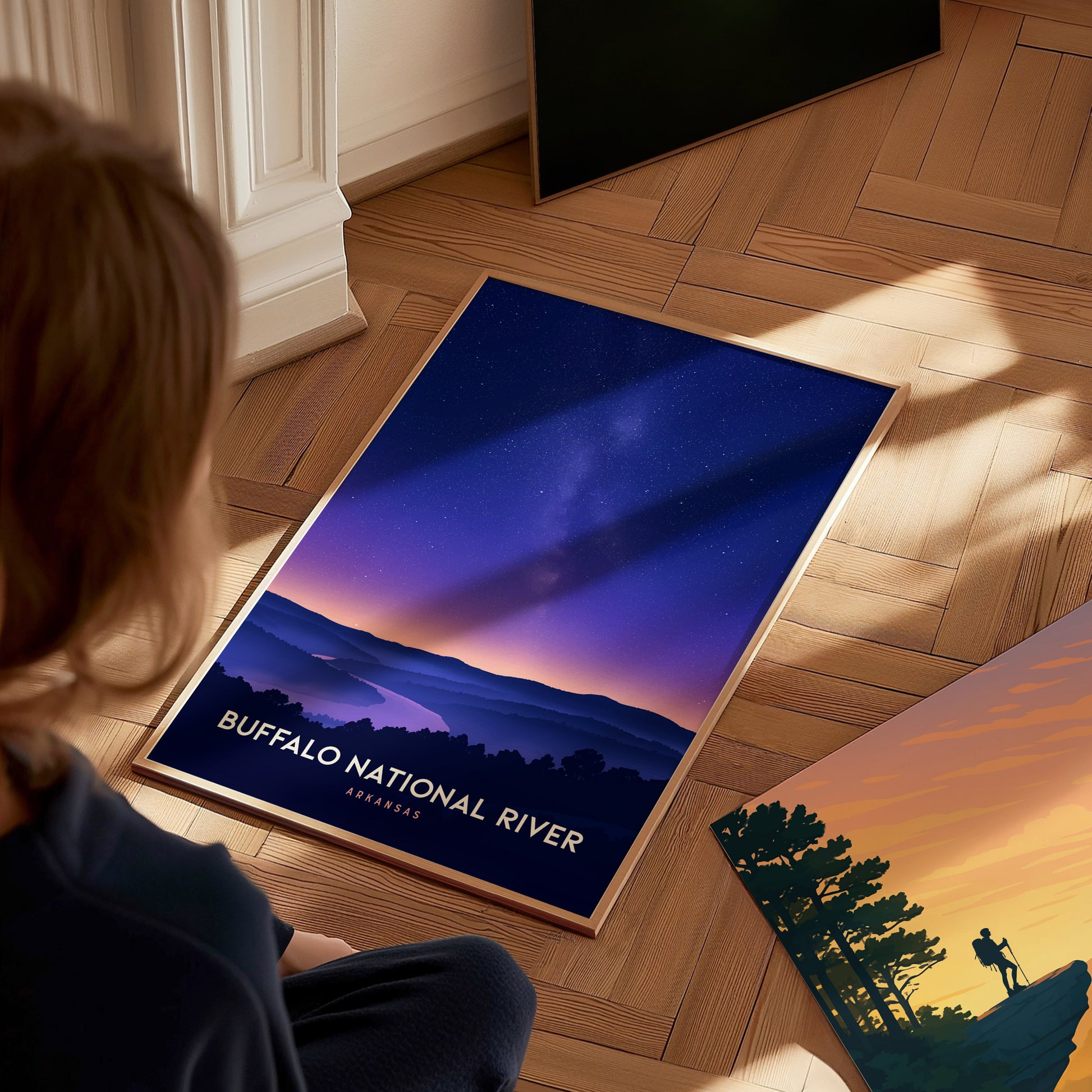 A child sits on a wooden floor, gazing at a framed travel print featuring the Buffalo National River. The artwork showcases a serene landscape under a starry sky, with mountains and trees silhouetted against a gradient of purple.