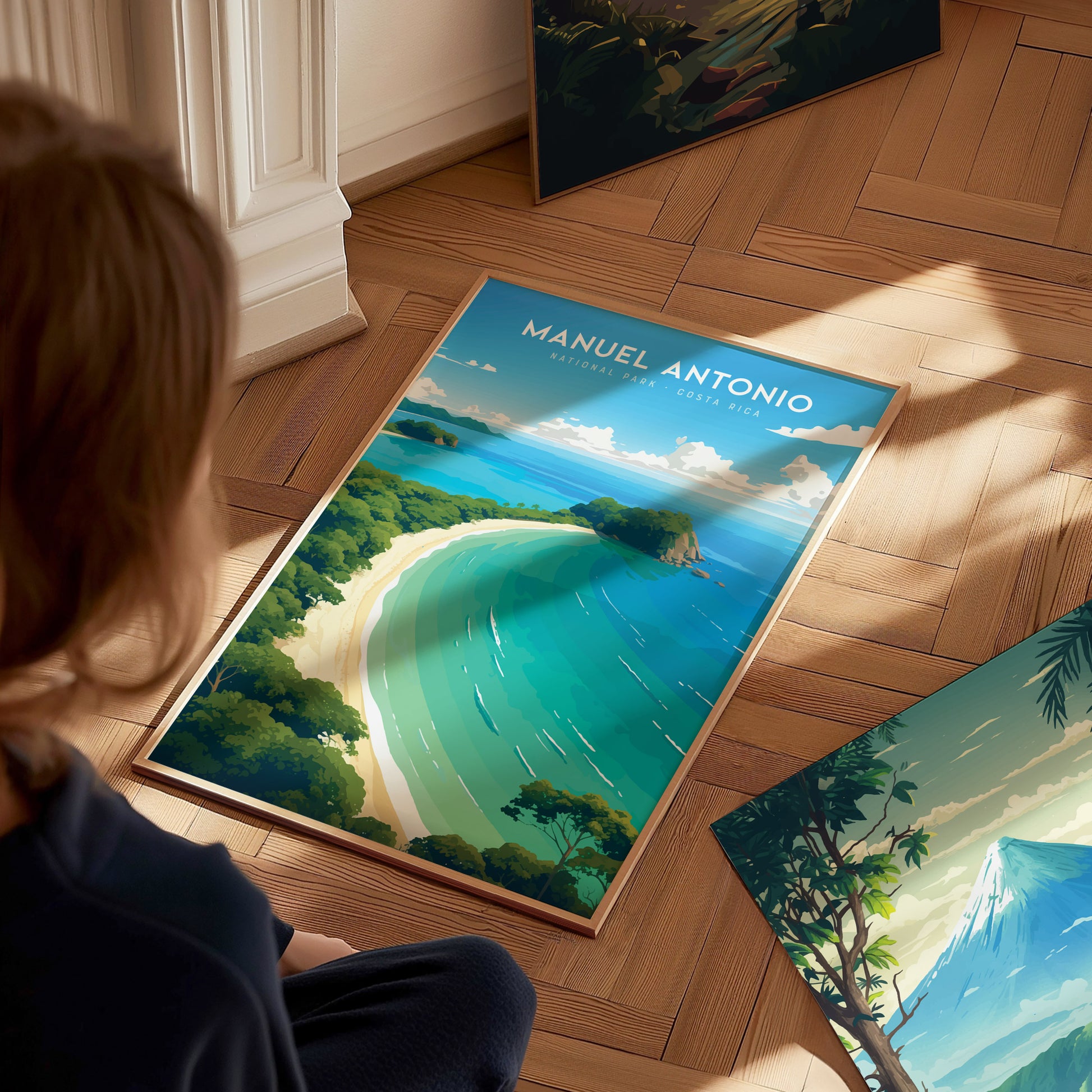 A person is sitting on a wooden floor, looking at a vibrant poster of Manuel Antonio National Park featuring turquoise waters, white sand, and lush greenery. Sunlight casts soft shadows.